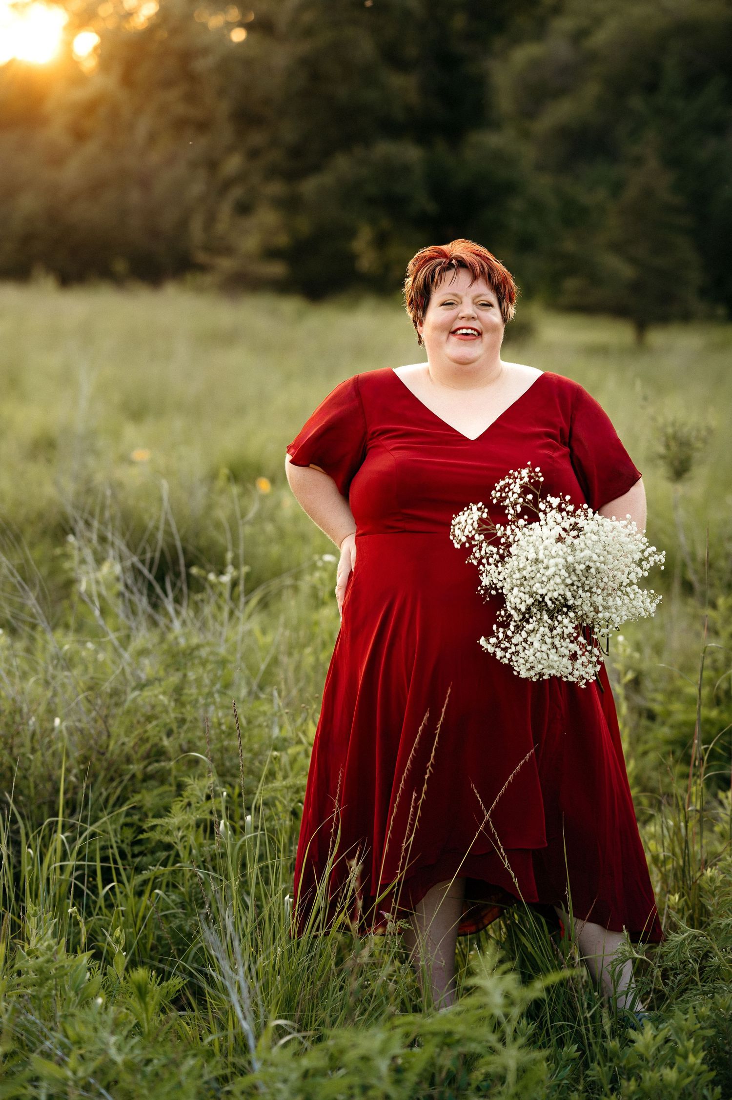 Adult woman in a red dress holding flowers in a green field with warm natural light and timeless Brookings SD senior photo styling.