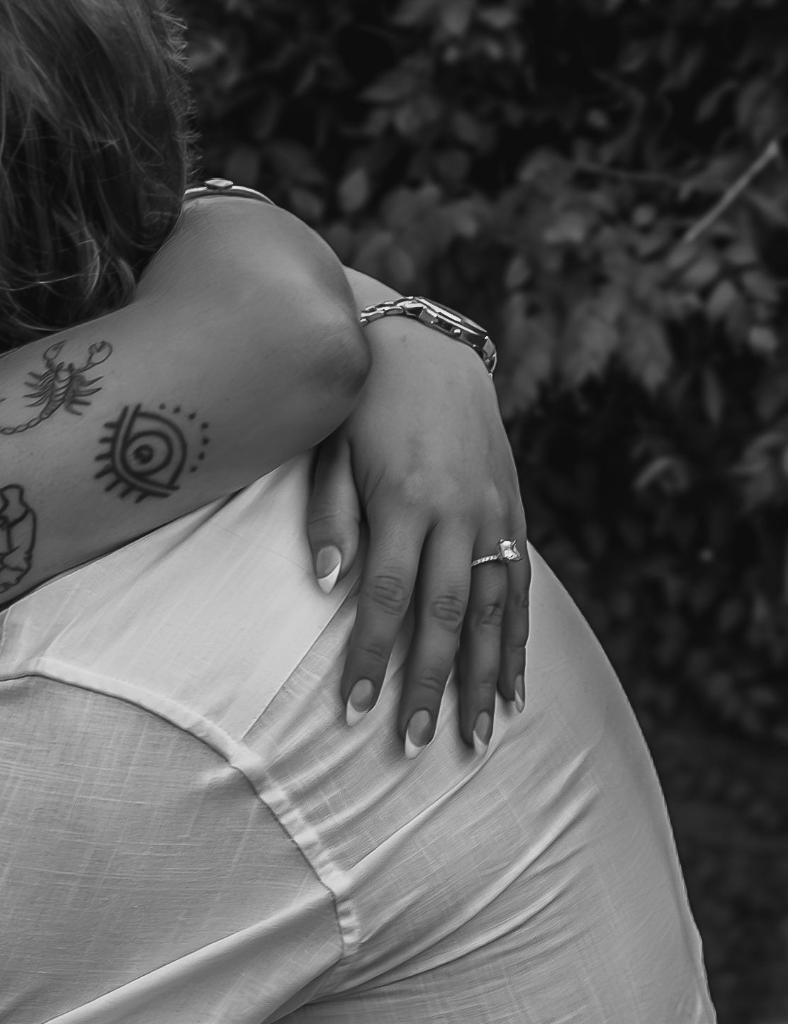 Black and white photo of a couple embracing, showing tattoos, a bracelet, and an engagement ring on the woman's hand.