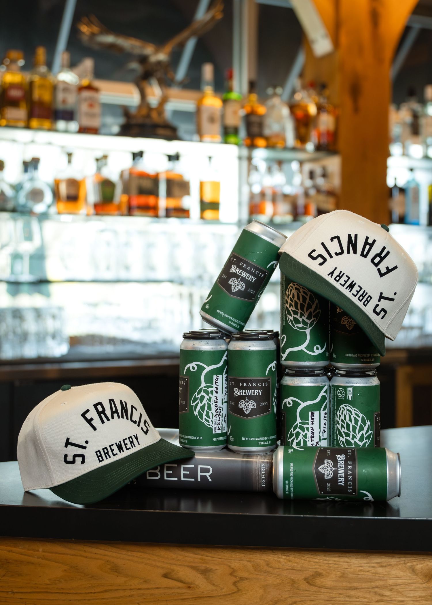 St. Francis Brewery Co. green beer cans stacked with branded white caps on a bar counter with liquor bottles in background.