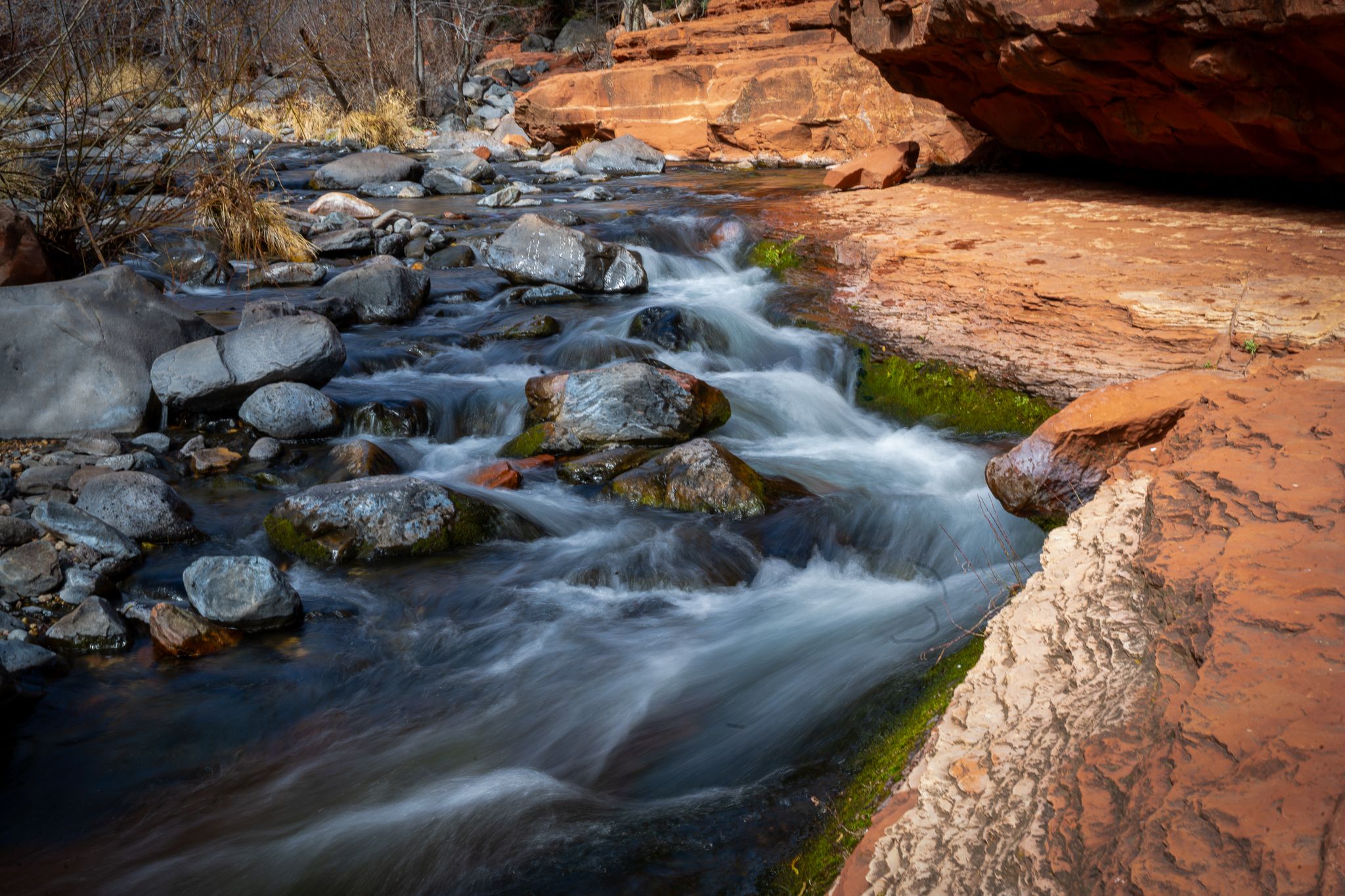 Long exposure of rushing stream flowing over rocks through red sandstone canyon walls.