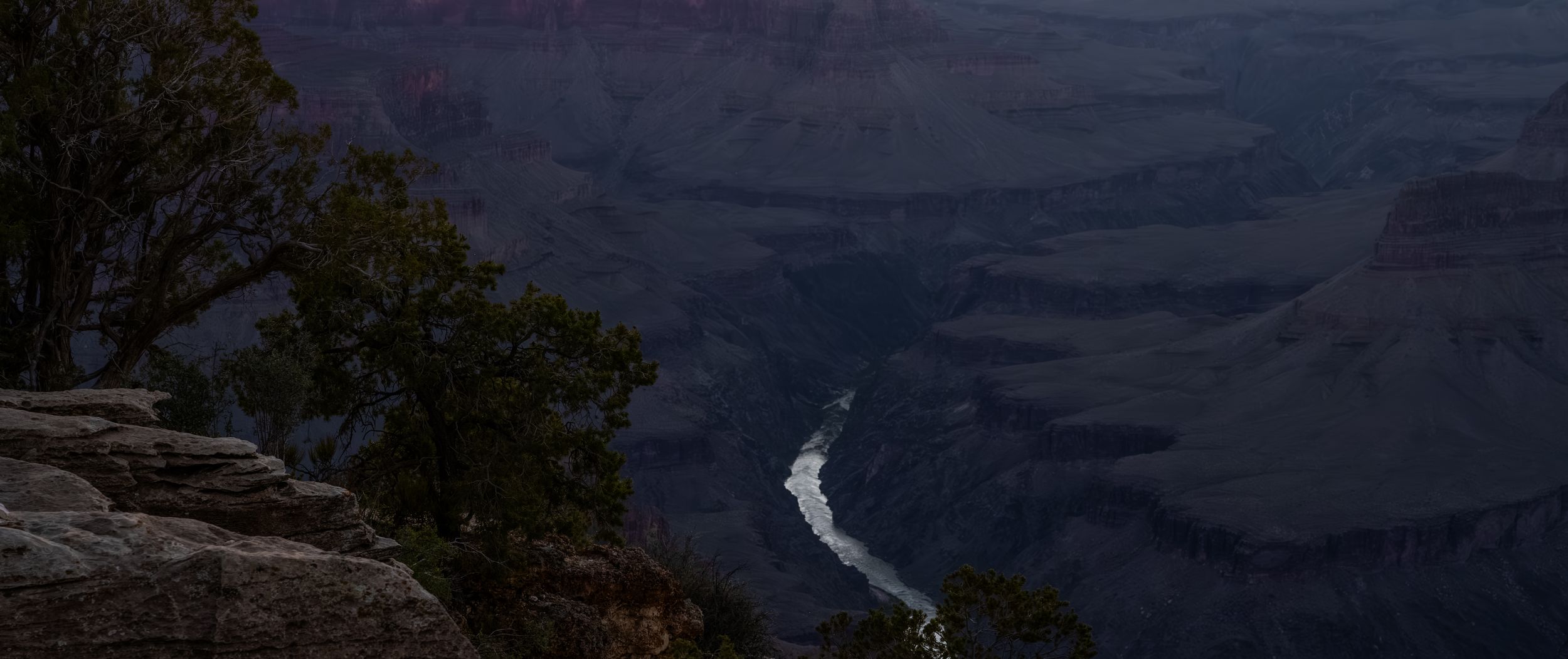 Twilight view of the Grand Canyon with the Colorado River reflecting light through shadowy layered rock formations.
