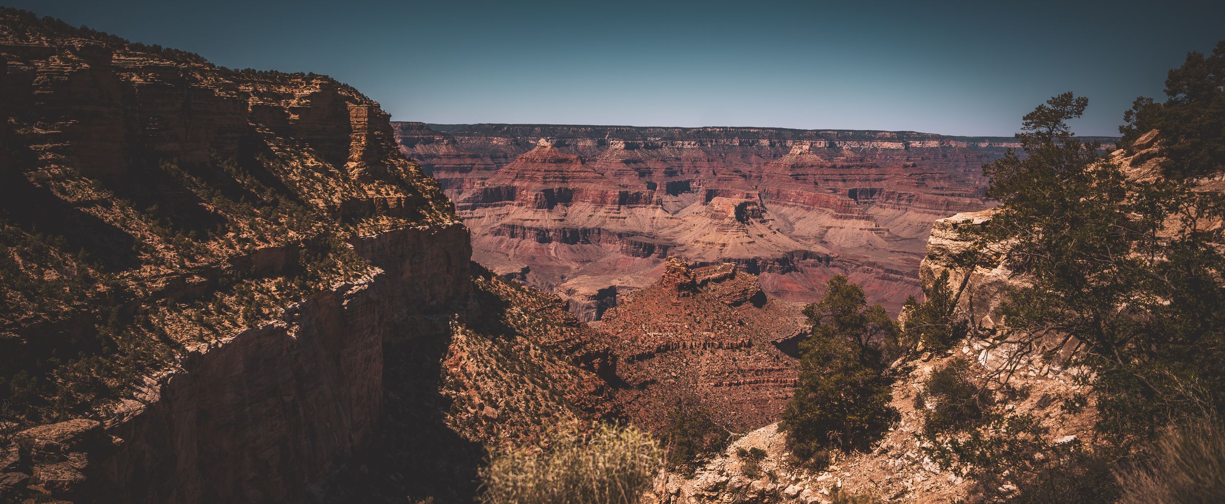 Panoramic aerial view of Grand Canyon's vast red rock formations and deep gorges under a clear blue sky.