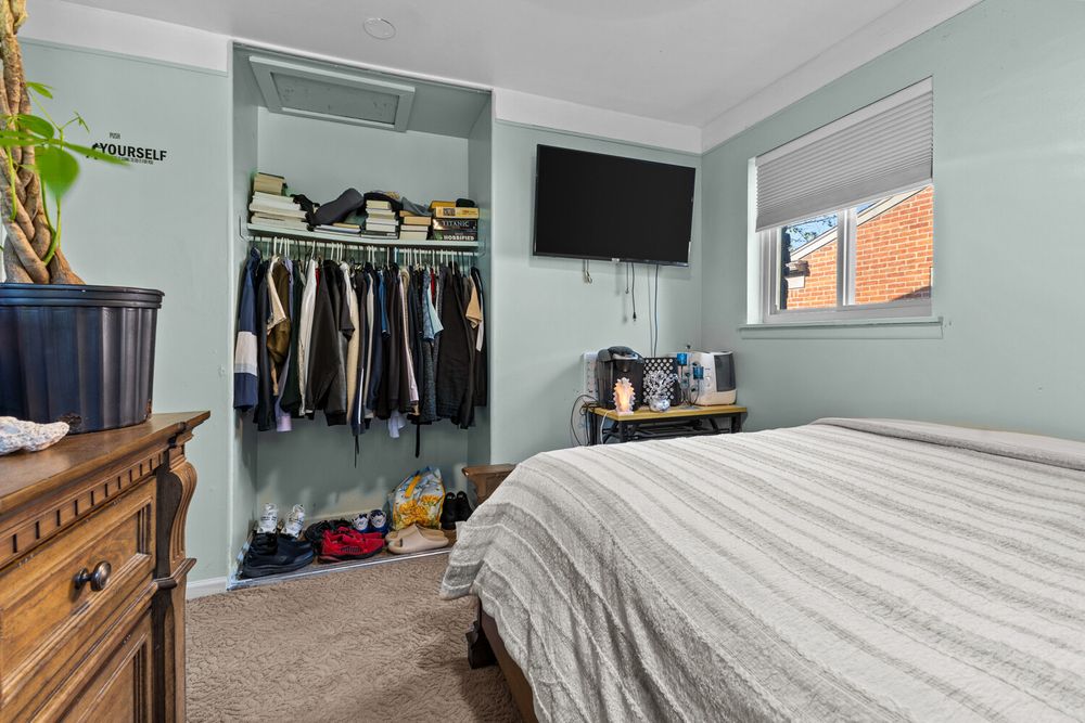 Bedroom with open closet full of clothing, wall-mounted TV, wooden dresser, and light blue walls with carpet.