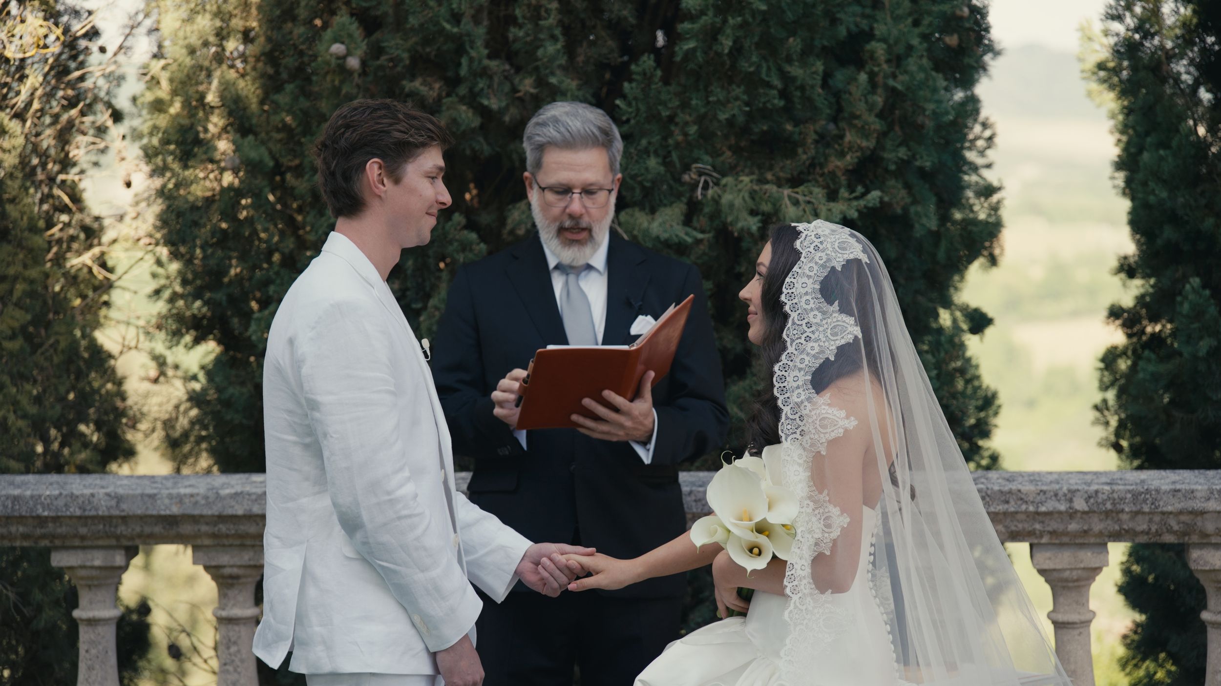 Elegant outdoor wedding ceremony with bride in white lace veil and groom in white suit, officiant reading vows.