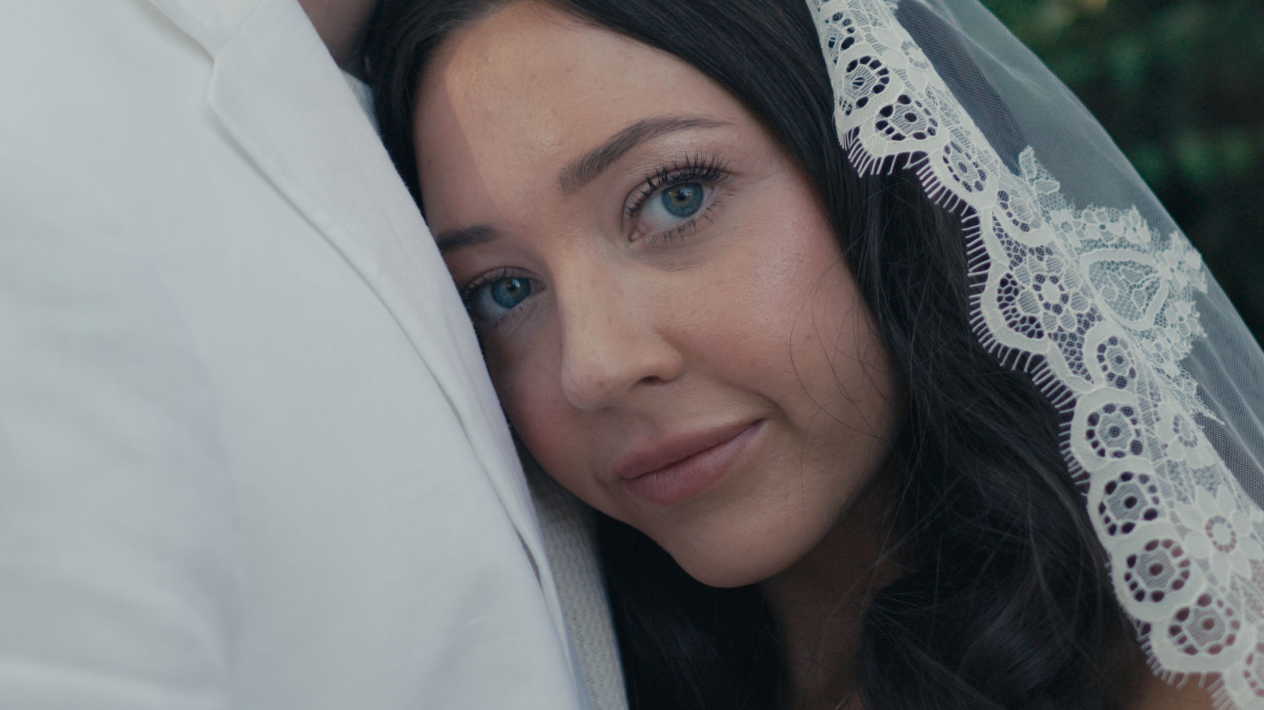 Bride in white veil with blue eyes leaning on groom's shoulder on wedding day.
