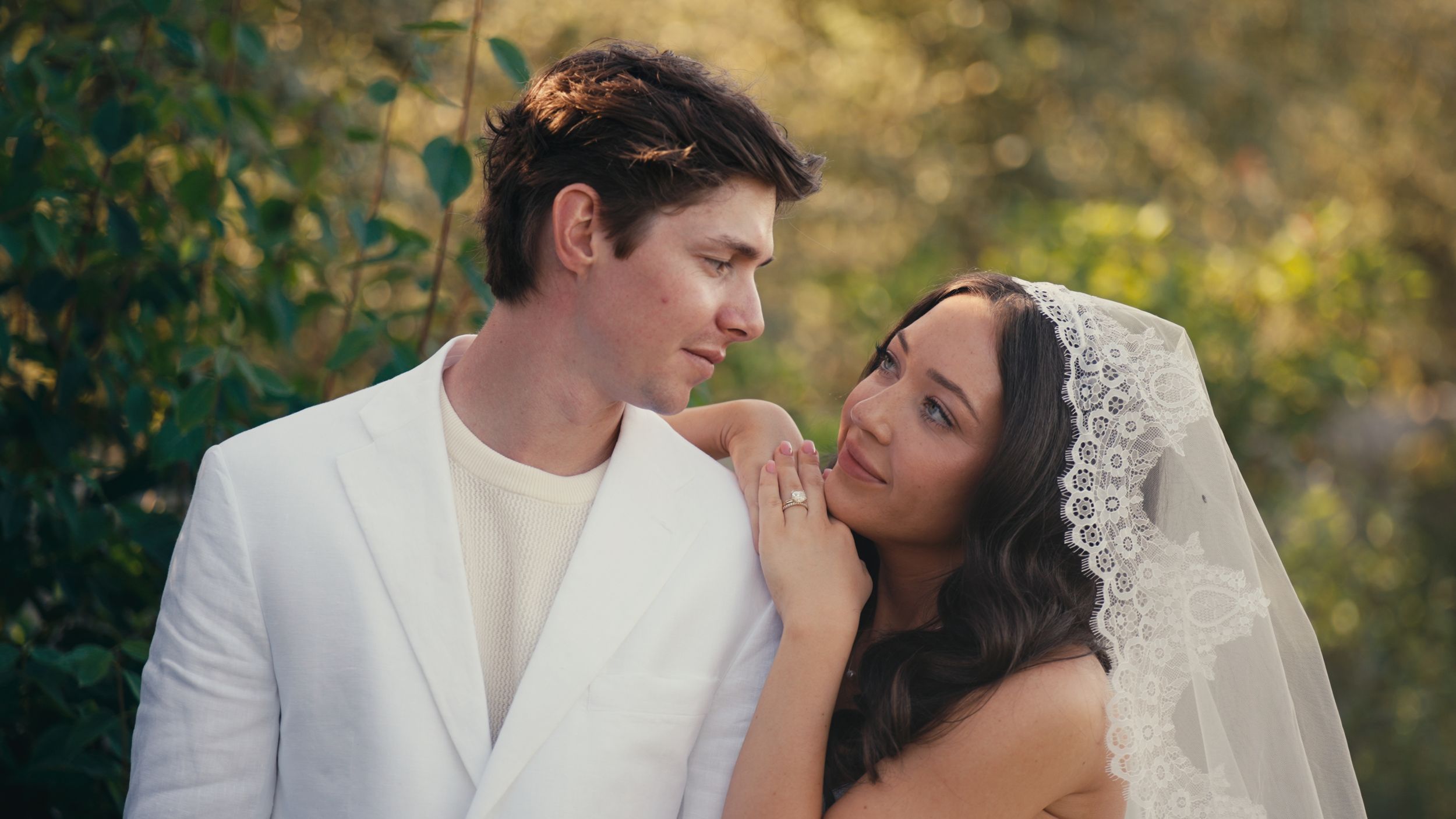 A couple in wedding attire sharing a romantic moment outdoors, groom in white suit, bride in lace veil.