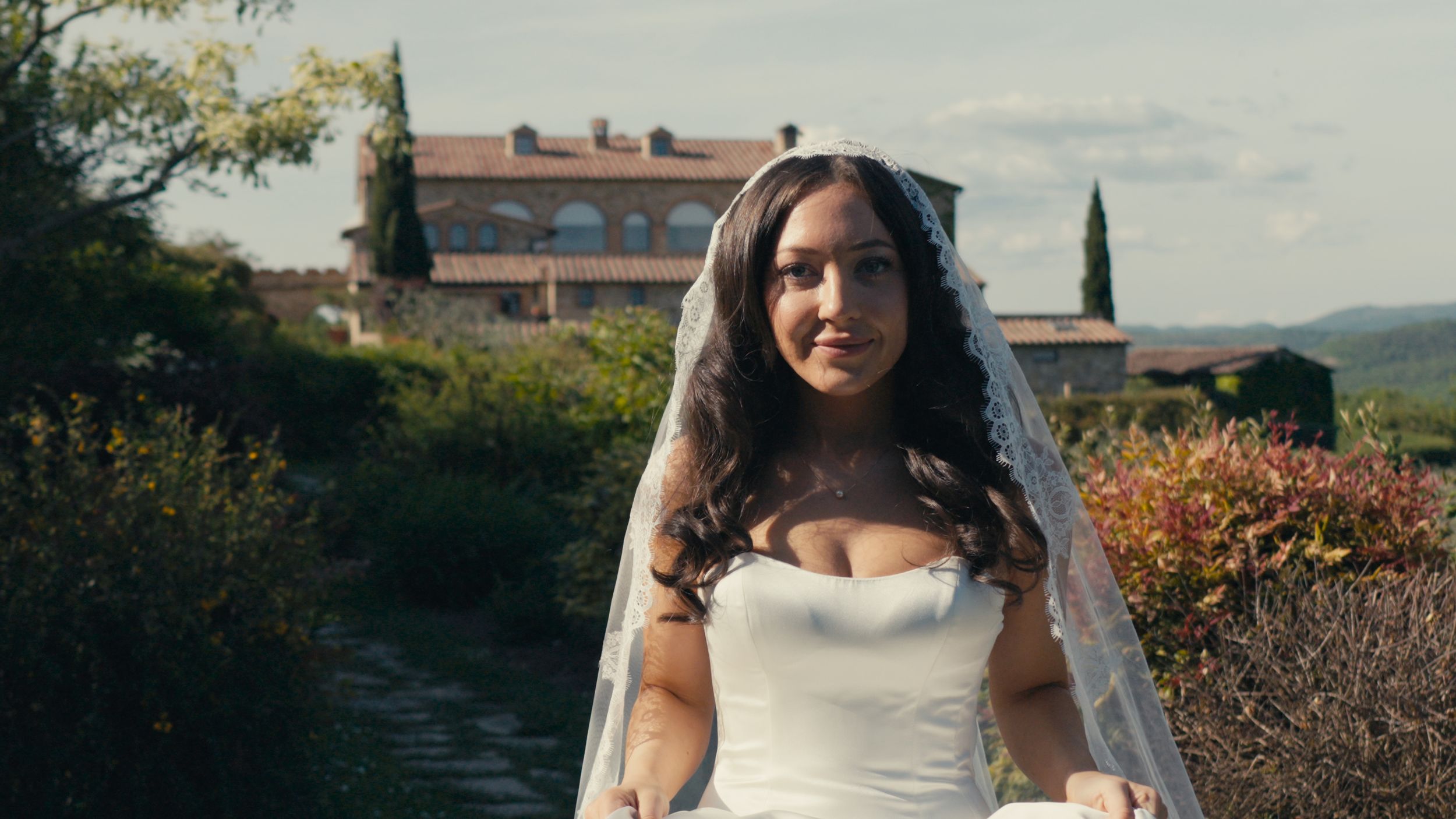 Bride in strapless white gown and veil walks along garden path at rustic venue.