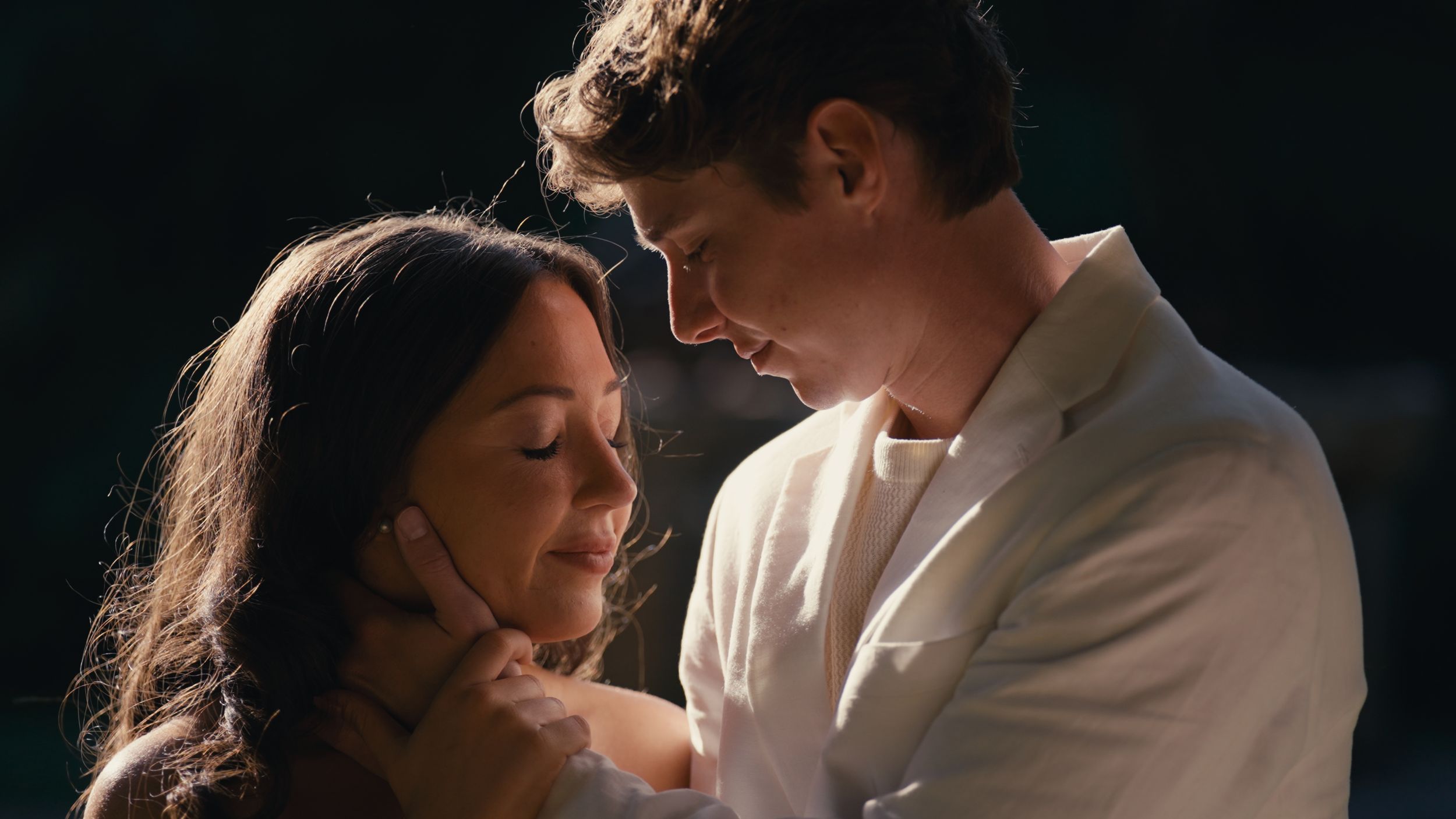 A romantic couple shares an intimate embrace outdoors, with a young woman and man in a white suit leaning close together.