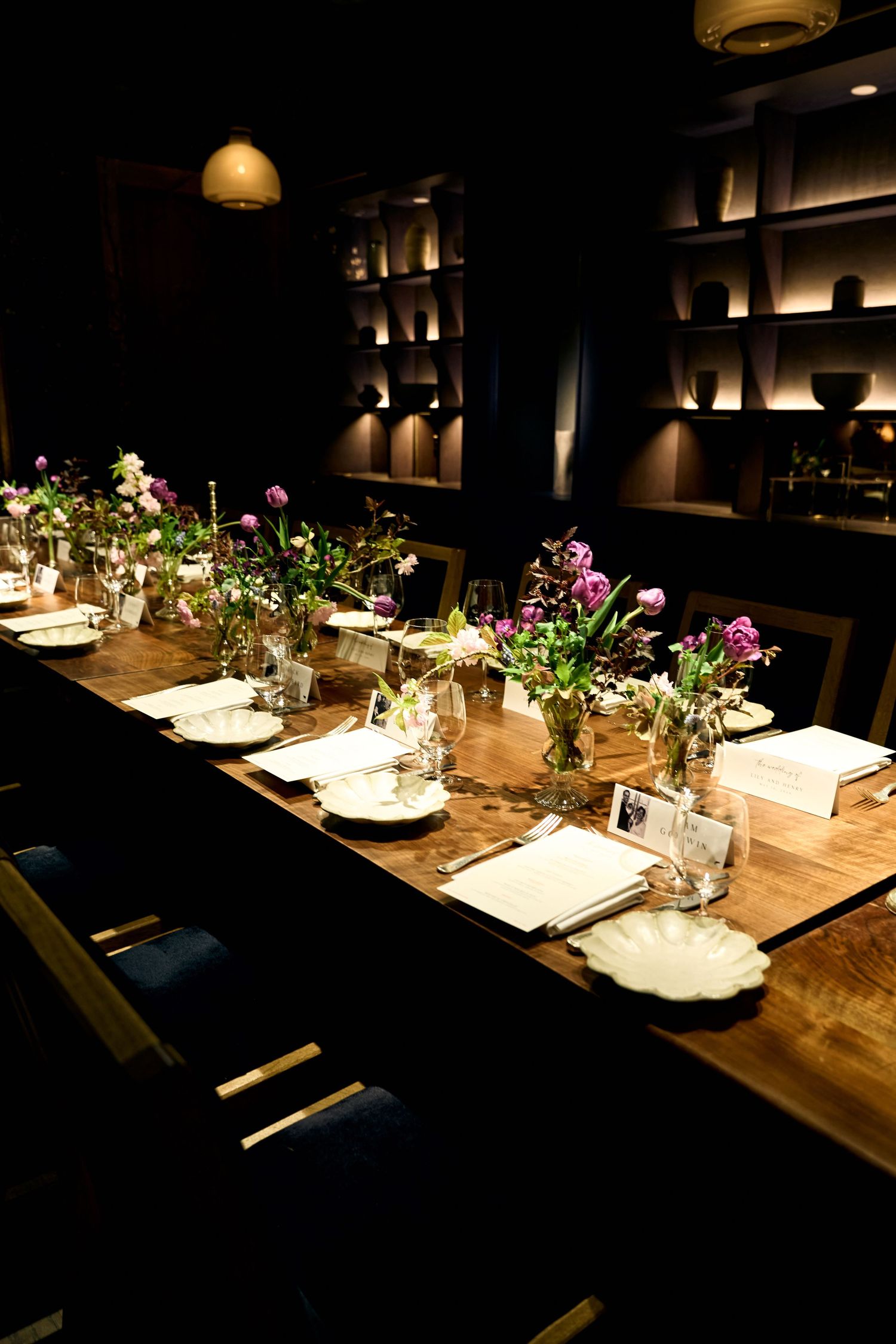 Elegant private dining room with long wooden table set with floral arrangements, white plates, and glassware in moody lighting.