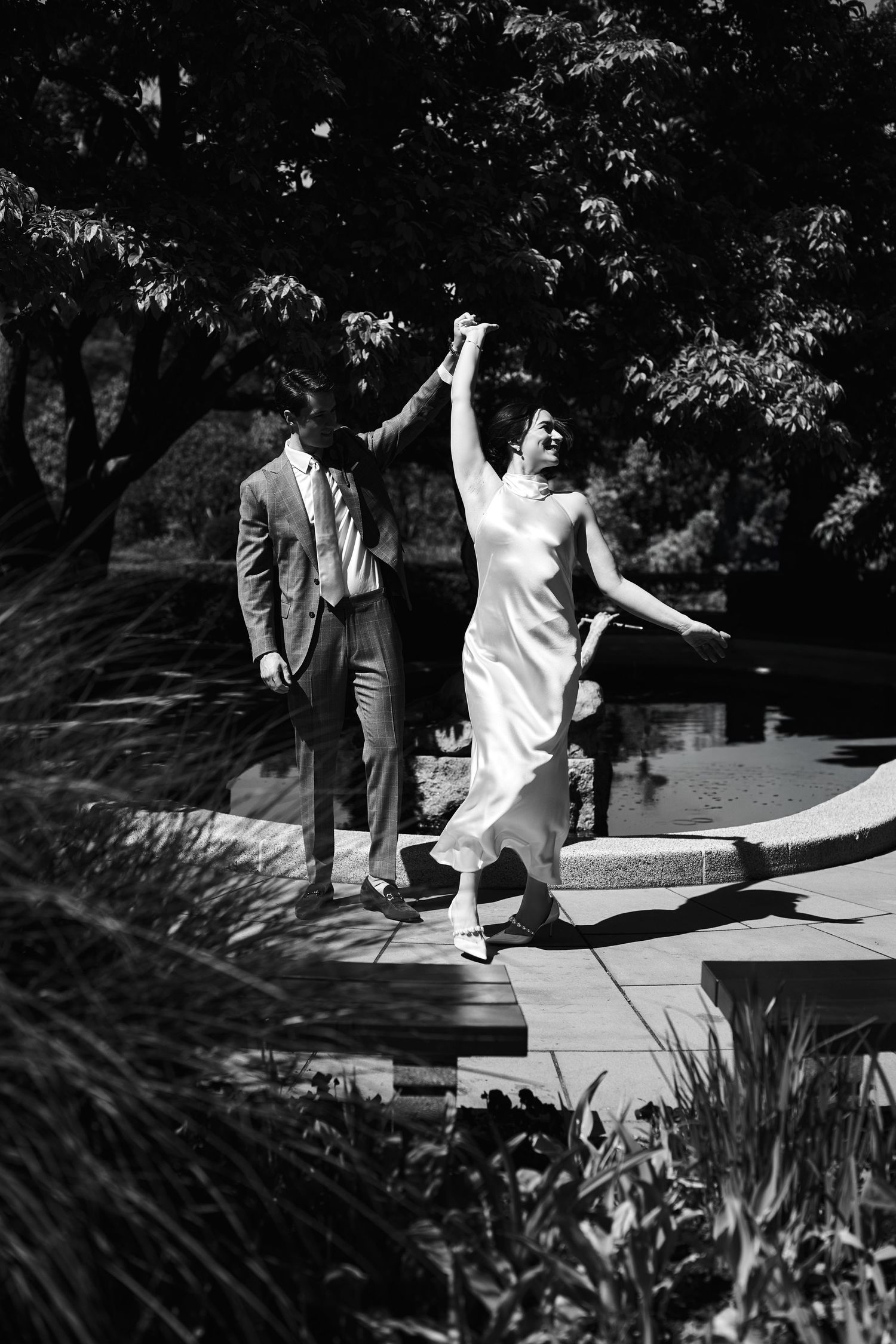 Black and white photo of a bride and groom dancing outdoors near a pond, surrounded by lush trees and greenery.