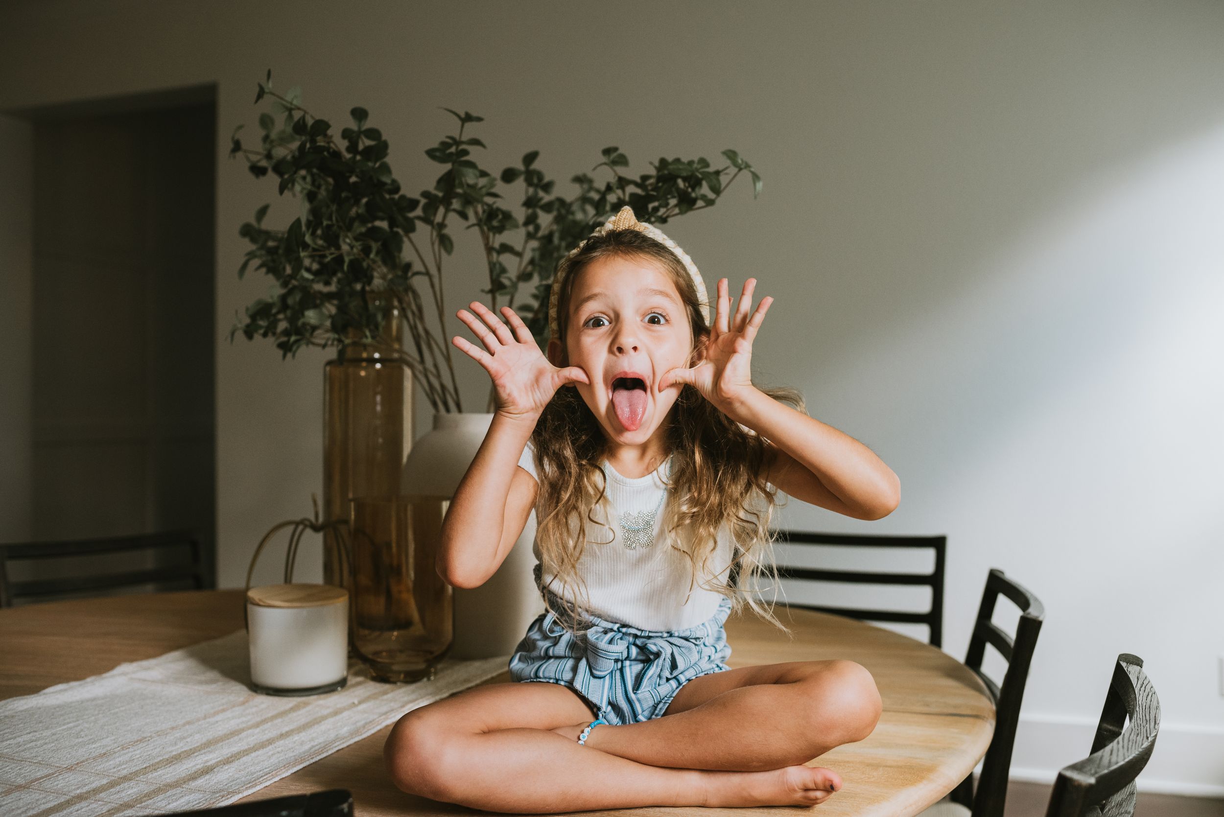 Cute little girl sitting cross-legged on table making funny face with hands up and tongue out indoors during an orlando photo shoot.
