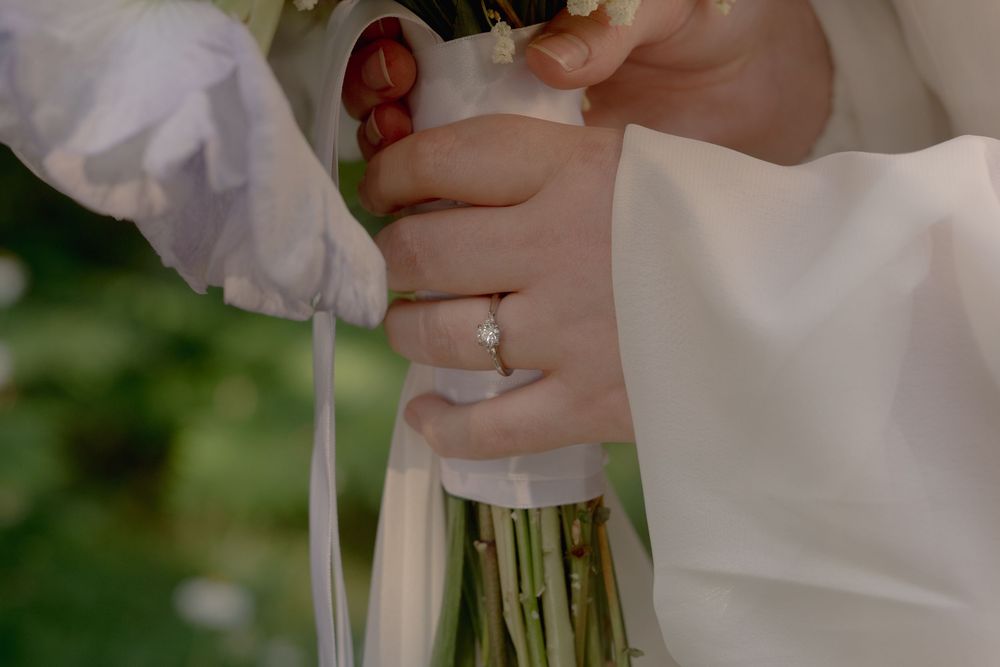 Bride holding a white wedding bouquet with a diamond engagement ring on her finger, wearing a white dress.
