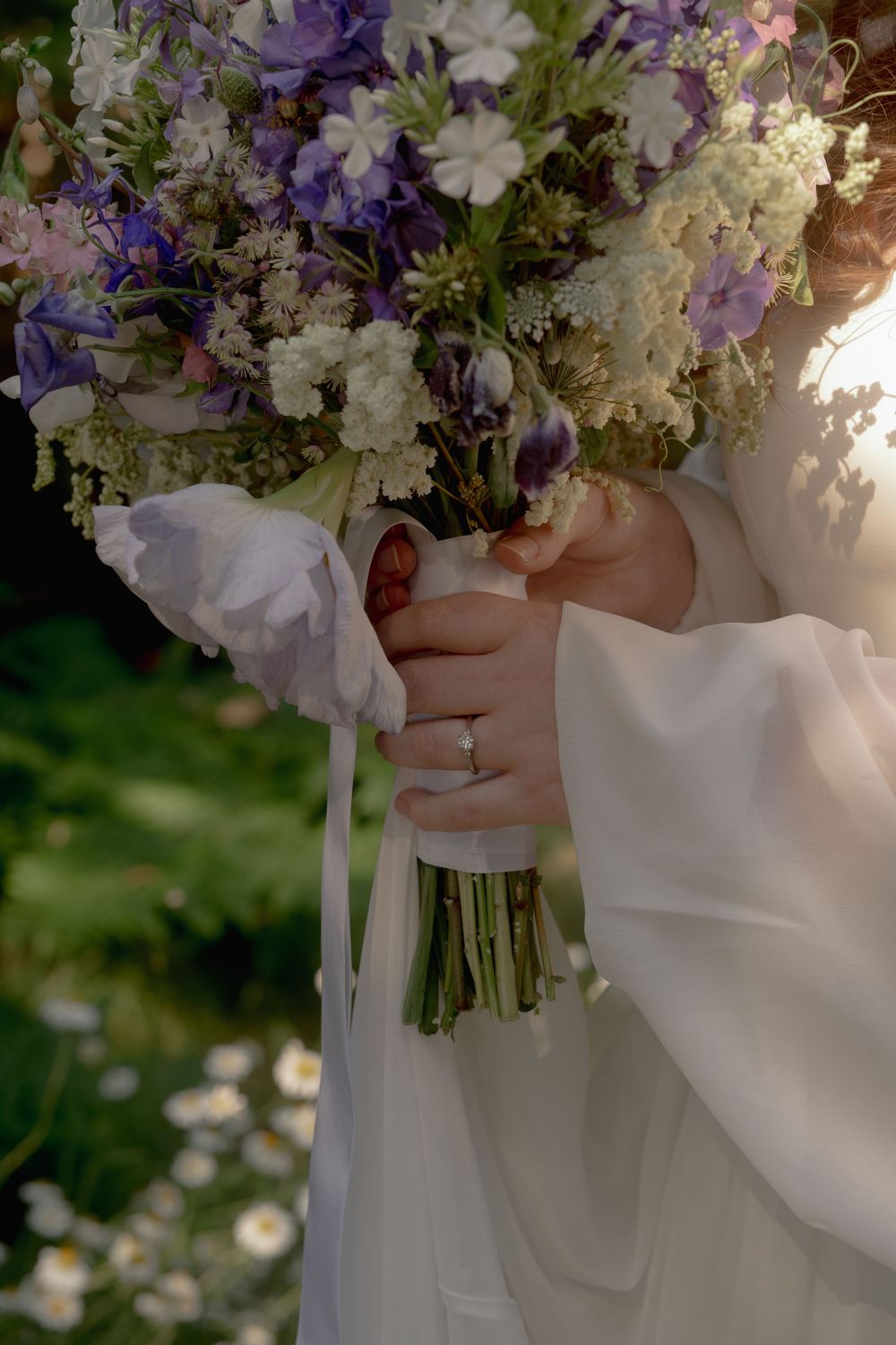 Bride holding a wildflower bouquet of purple and white blooms with ribbon, wearing a white gown and diamond ring.