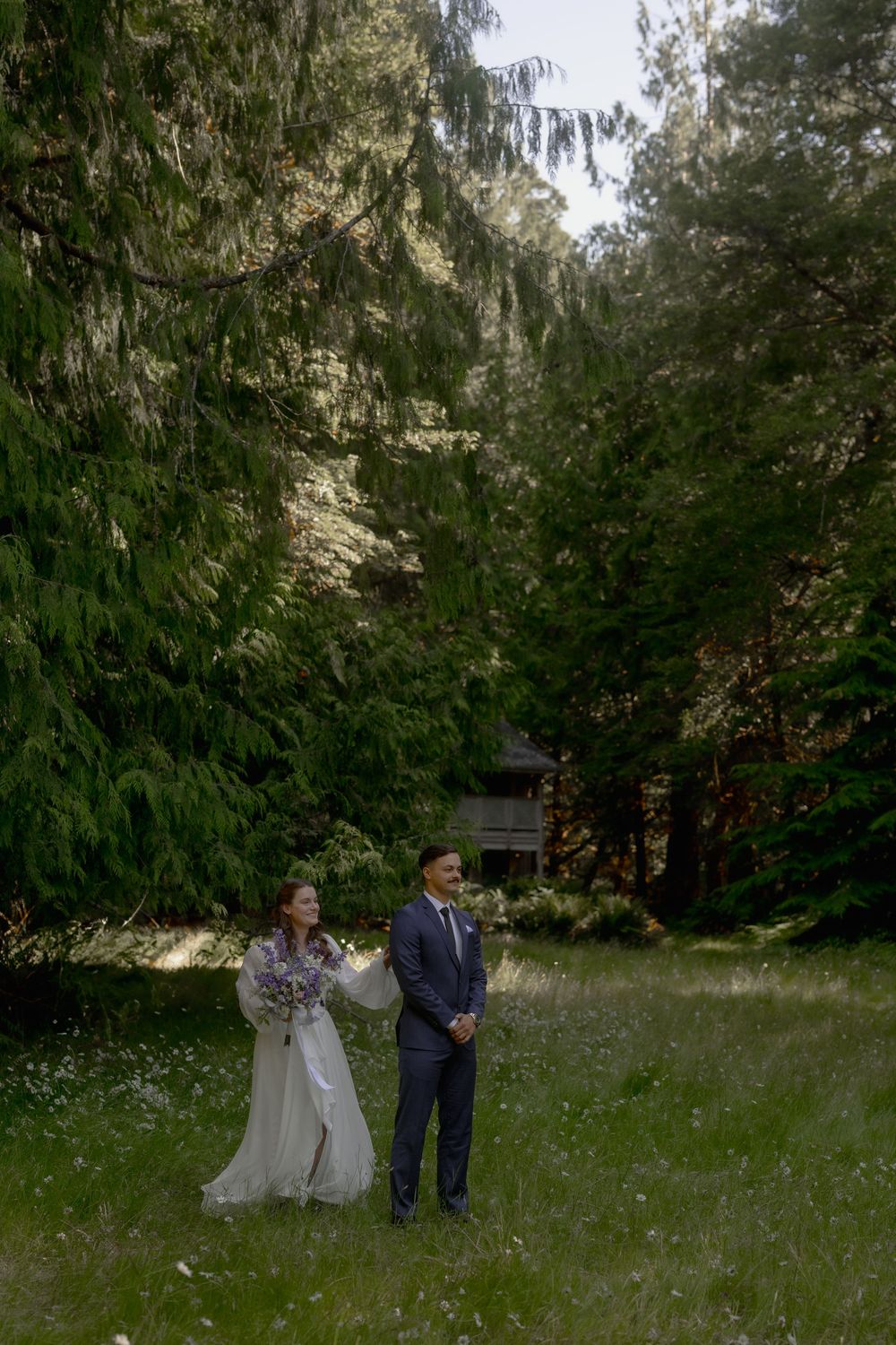 Bride and groom stand in a lush green forest meadow during their outdoor wedding photo session.