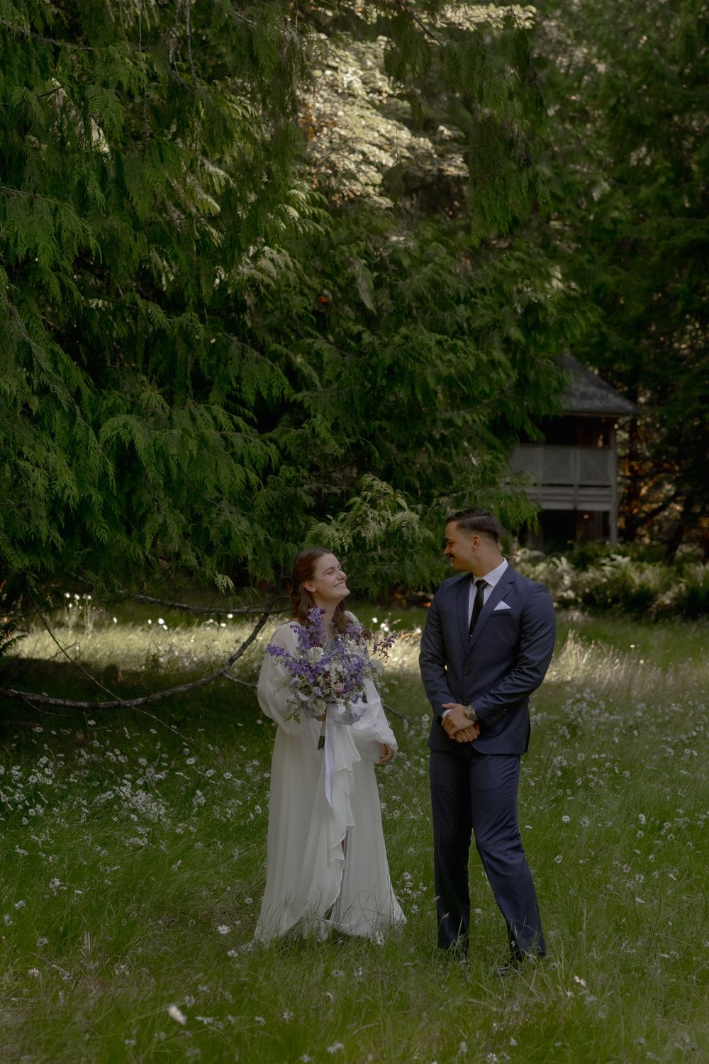 Bride and groom share a joyful moment in a lush green meadow, bride holding a purple wildflower bouquet.