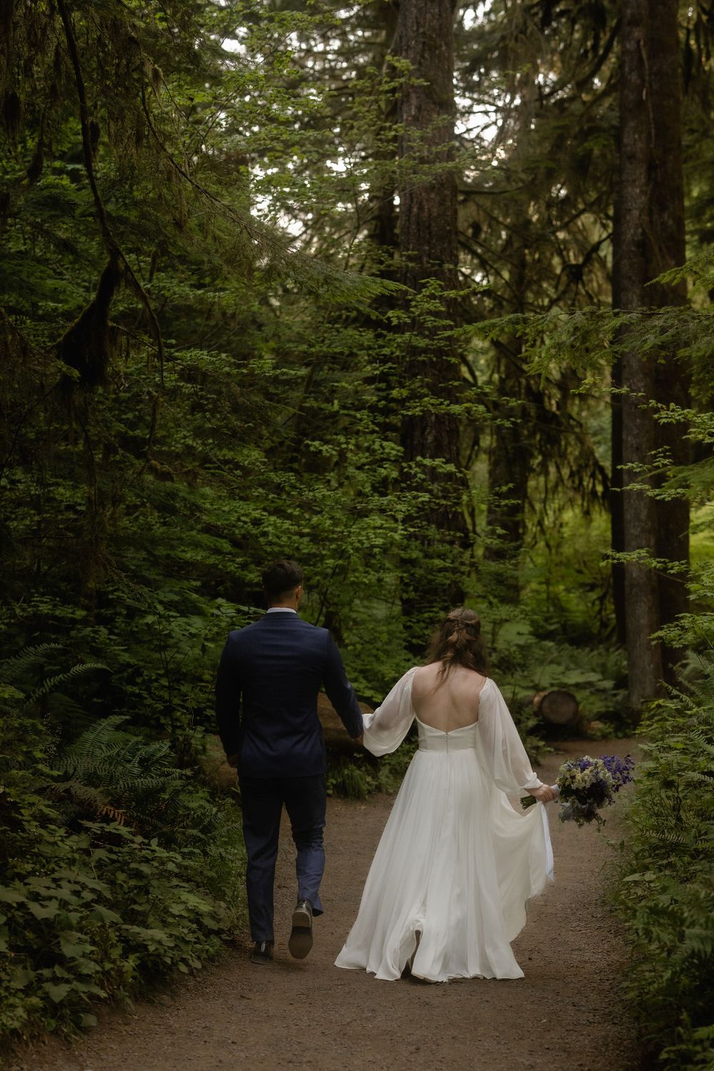 Bride and groom walking hand in hand through a lush green forest on their wedding day.