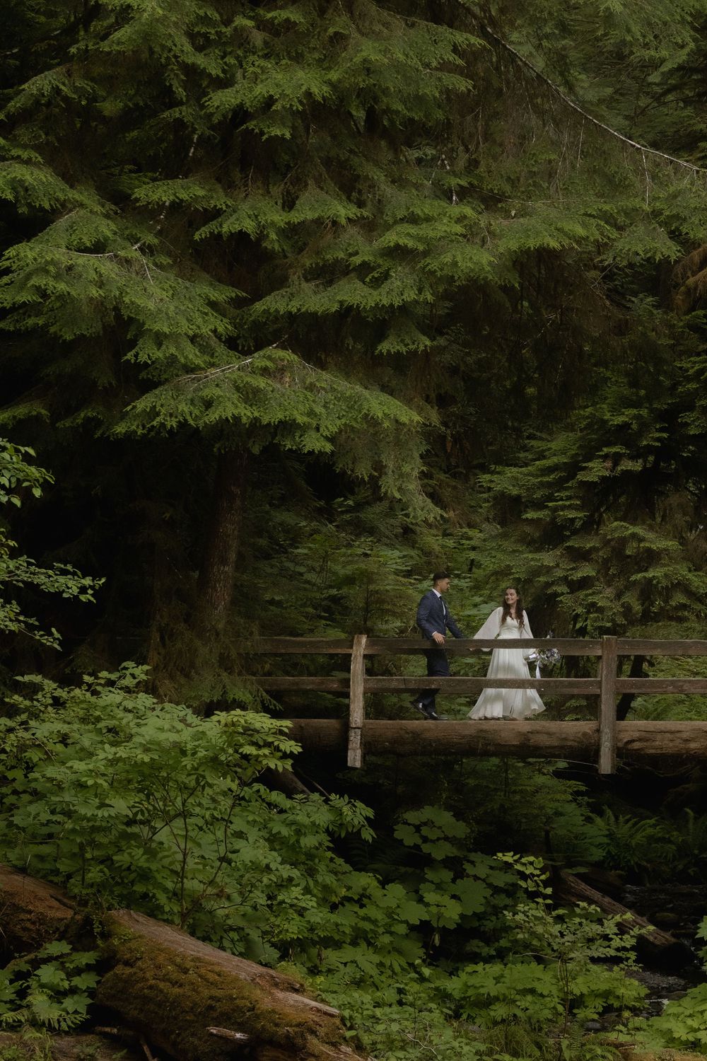 A bride and groom walk hand in hand across a wooden bridge surrounded by lush green forest trees.