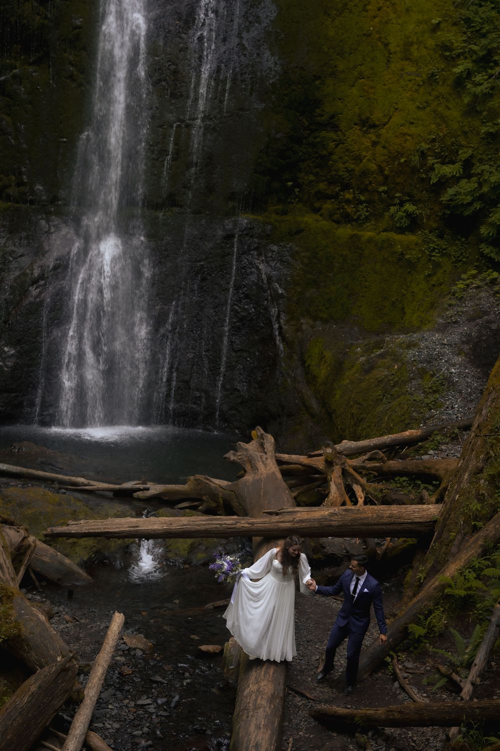 Bride and groom walk across logs in front of a stunning waterfall on their wedding day.
