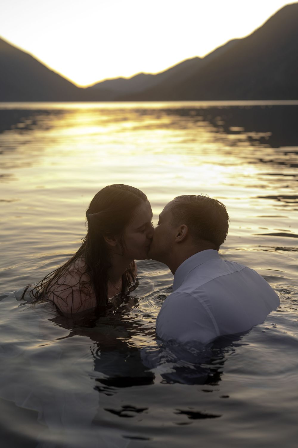 Couple kissing in a lake at golden sunset with mountain silhouettes in the background.