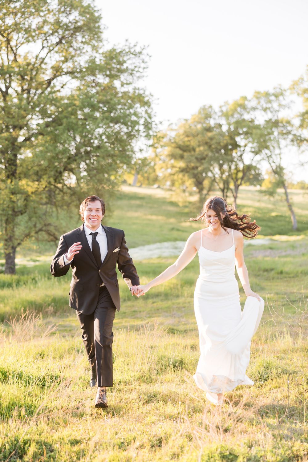 Bride and groom holding hands running through a sunlit green meadow, bride in white gown, groom in black tuxedo.