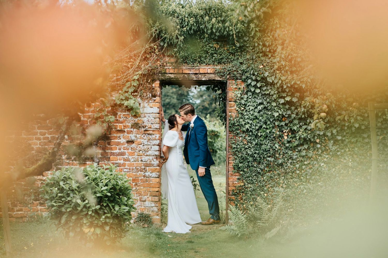 Bride and groom share a romantic kiss framed by an ivy-covered brick doorway surrounded by lush greenery.