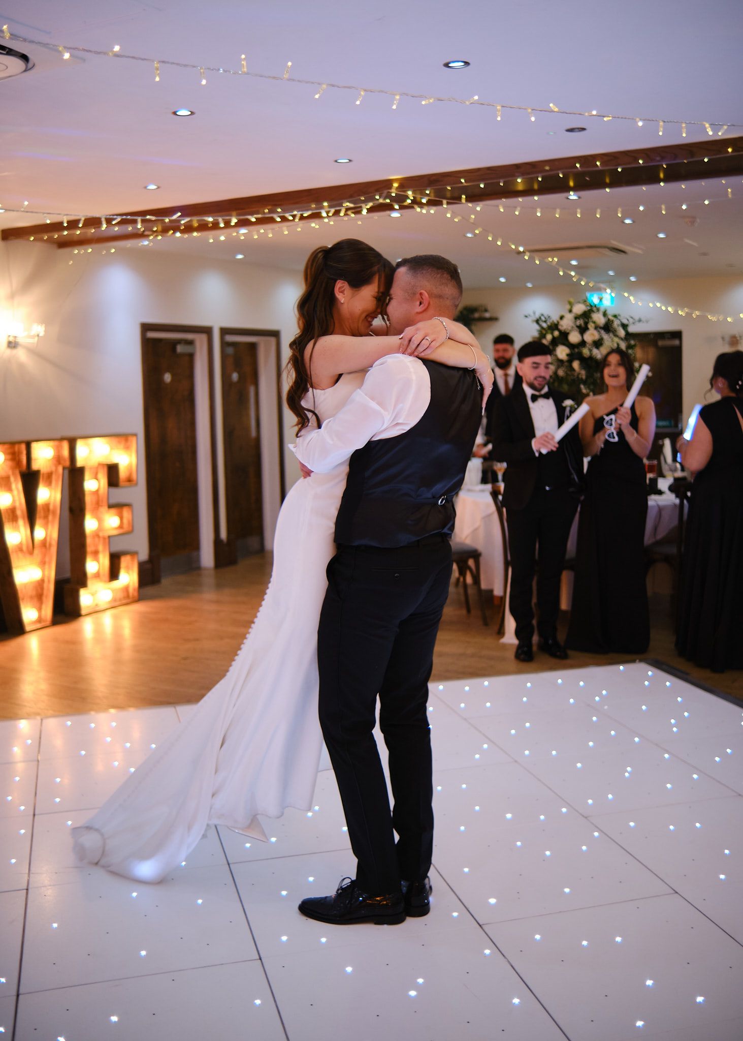 Bride and groom sharing their first dance on a white sparkle floor beside a glowing LOVE sign.