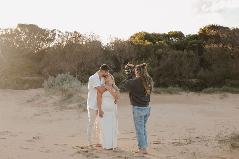 Photographer capturing couple embracing on sandy beach at golden hour.