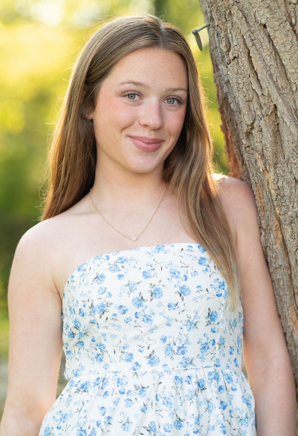 Young woman with long brown hair smiling in a white floral dress, leaning against a tree outdoors.