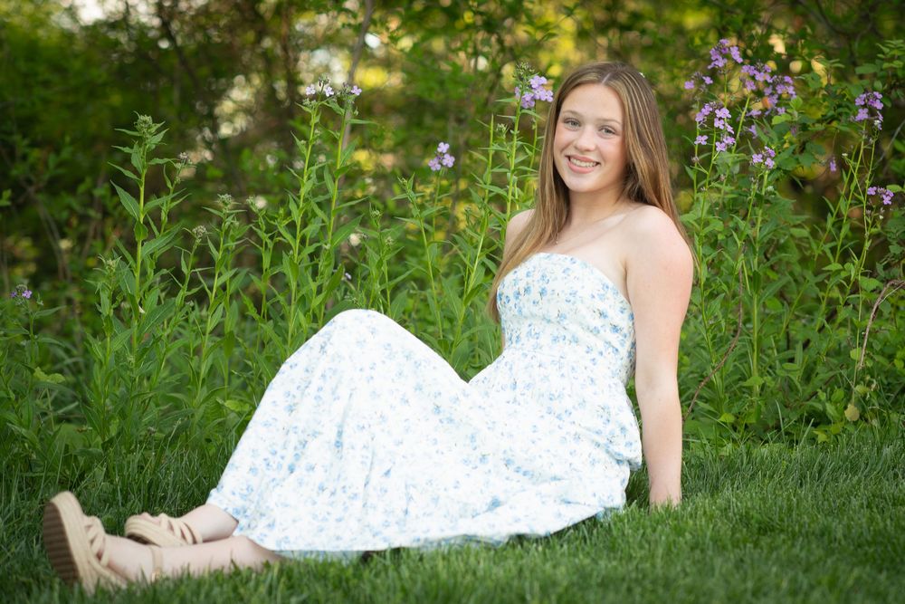 Young woman in floral sundress sitting on green grass surrounded by purple wildflowers in a garden setting.