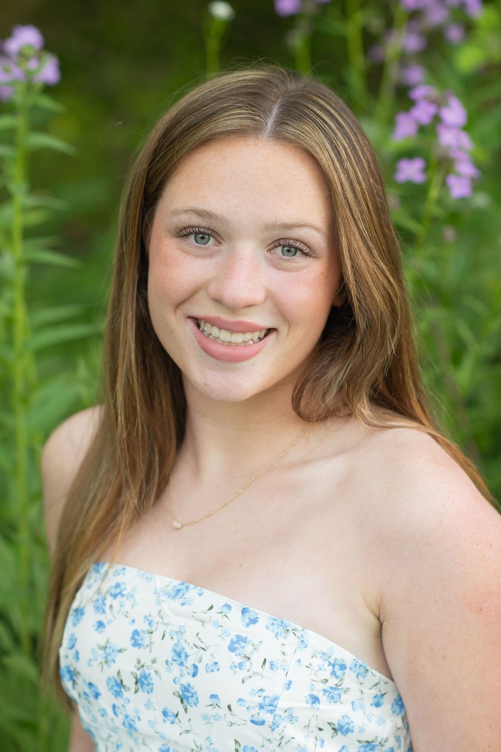 Smiling teenage girl with braces and long brown hair wearing a floral dress outdoors with purple wildflowers.