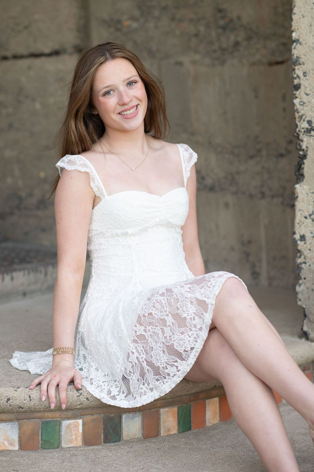 Young woman in white lace dress sitting on tiled steps, smiling outdoors.