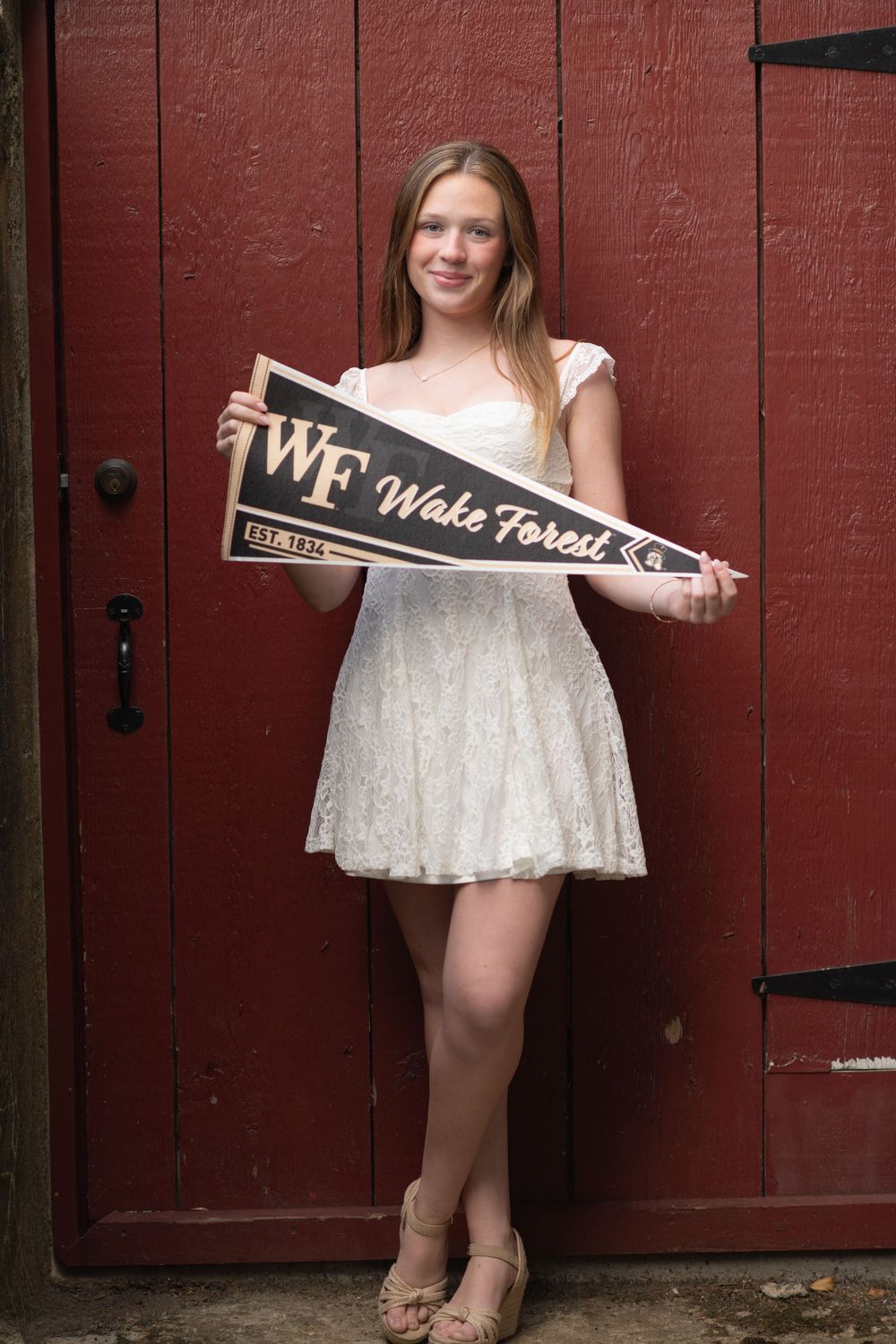 Young woman in white dress holding Wake Forest University pennant flag, posing in front of red barn doors.