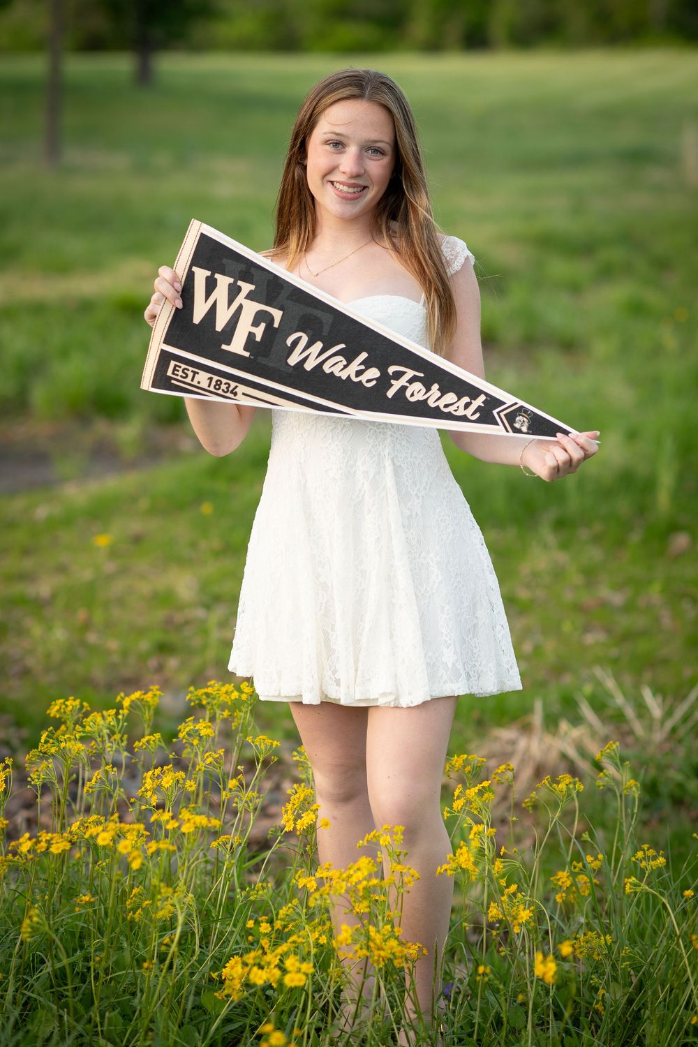 Smiling girl in white dress holding Wake Forest pennant flag in a field of yellow wildflowers.