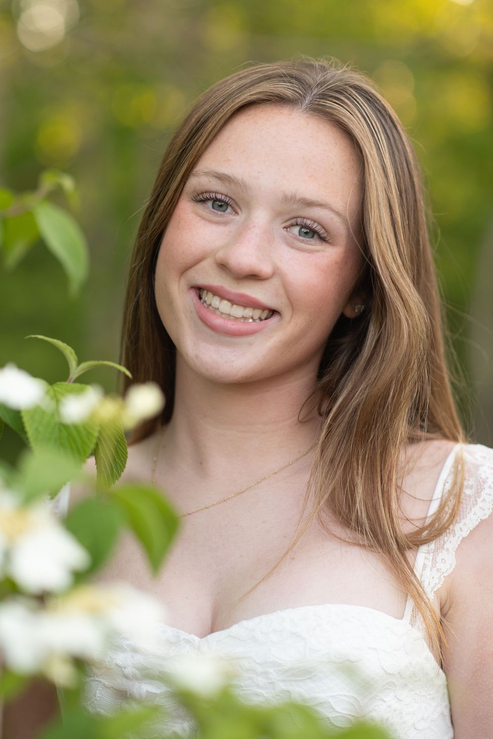 Smiling teenage girl with freckles and long brown hair wearing a white lace dress outdoors among green leaves and white flowers.