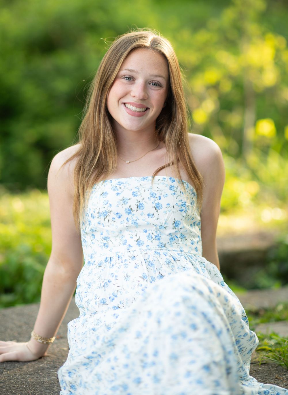 Young woman in a white floral sundress smiling outdoors with lush green foliage in the background.