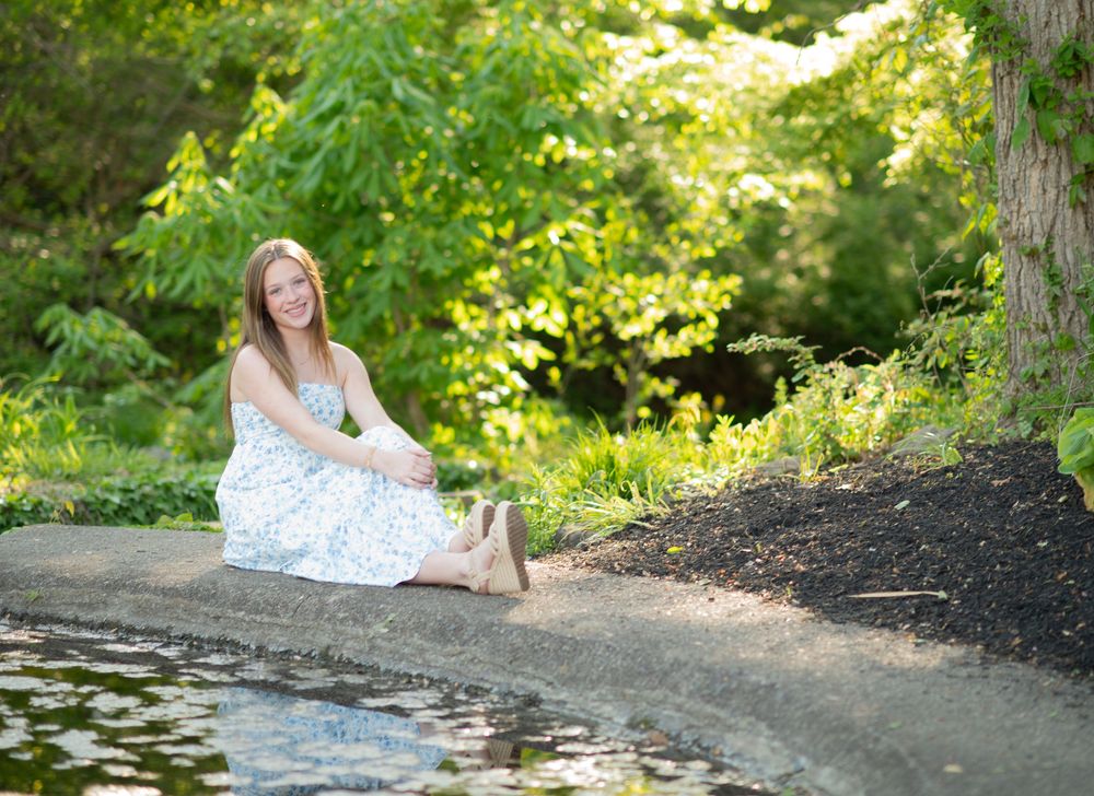 Smiling young woman in floral dress sitting barefoot by a garden pond surrounded by lush green trees.