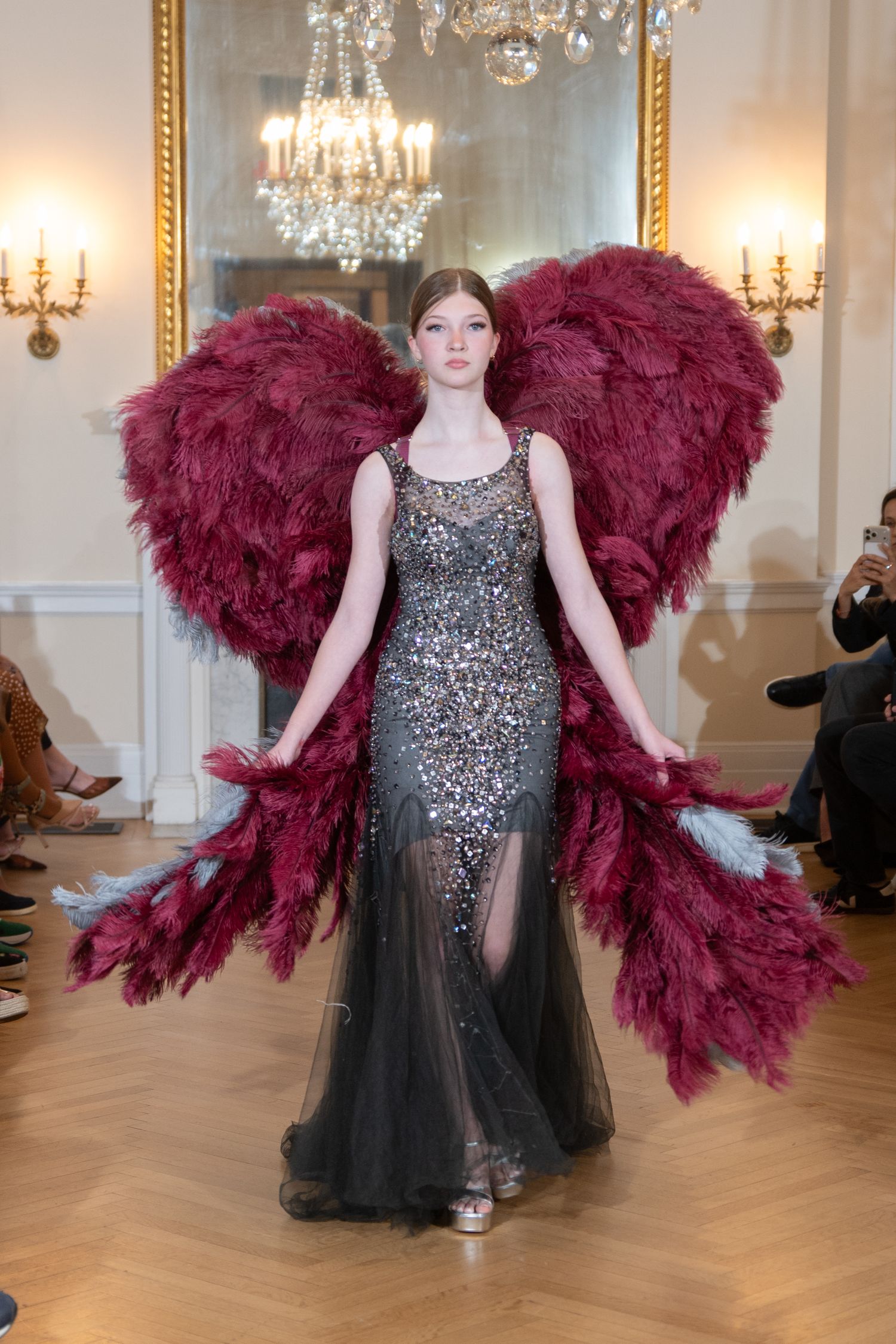 Model wearing a beaded gray gown with dramatic burgundy feather wings at a luxury fashion runway show.