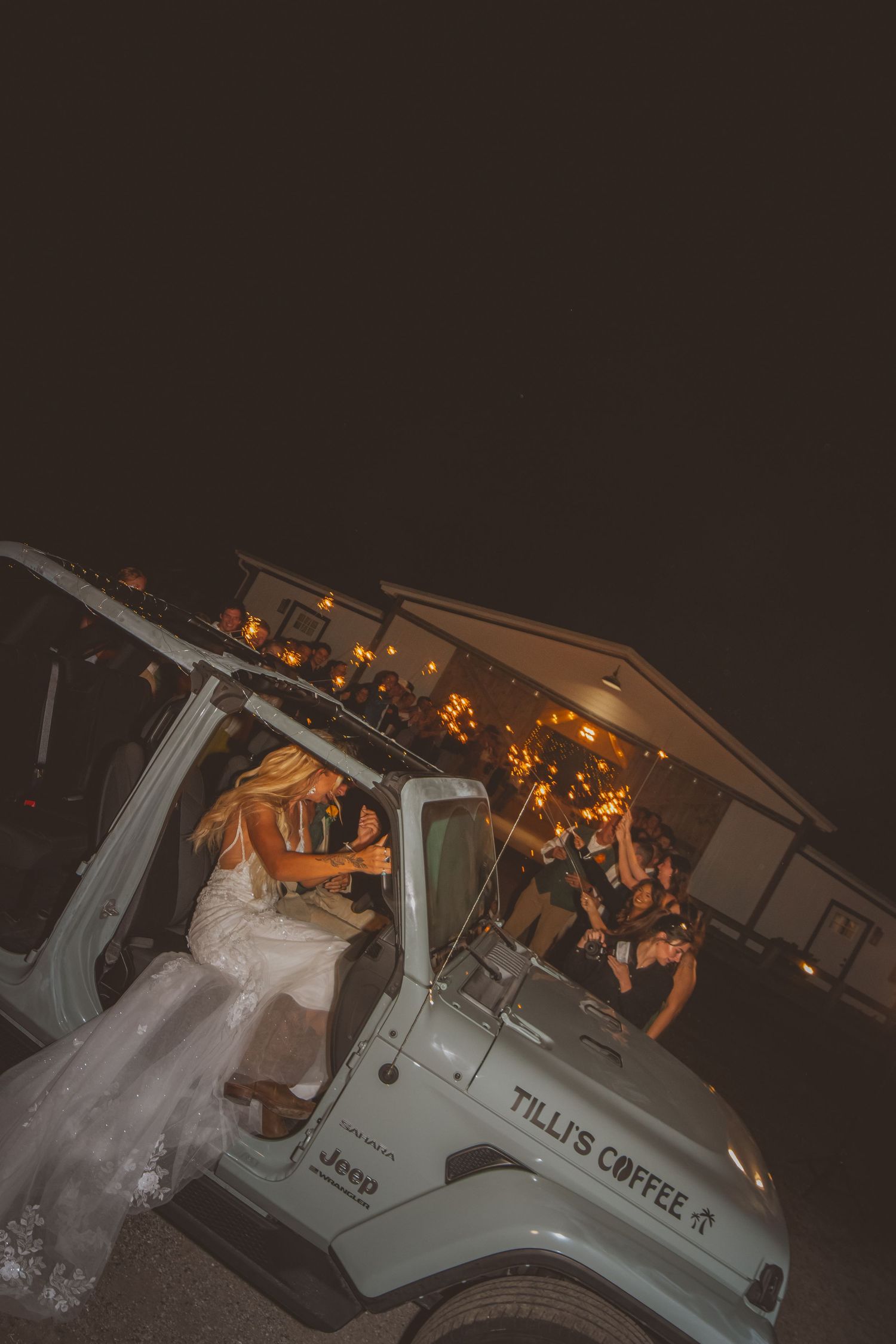 Bride, Tilli, gets into her blue Jeep in a wedding dress with the words "Tilli's Coffee" on the hood during a wedding sparkler sendoff