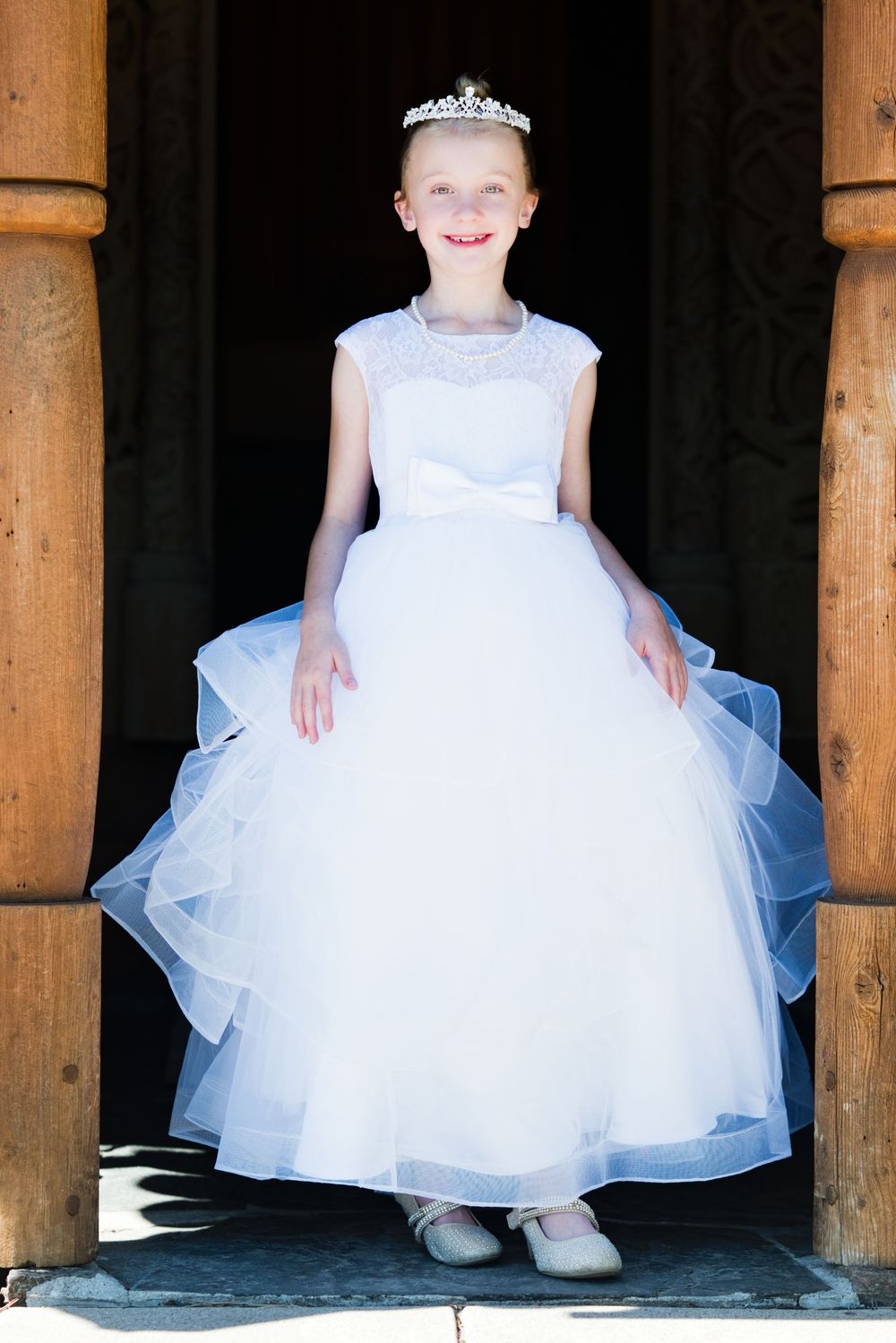 Young girl in white tulle dress and tiara stands smiling in wooden doorway for First Communion.
