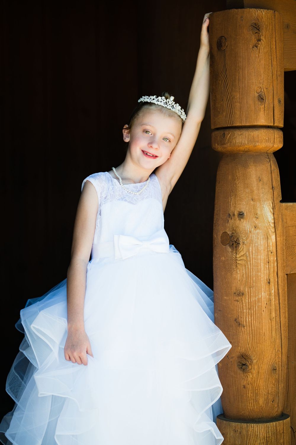 Young girl in white tiara and ruffled ball gown dress poses by wooden staircase post.