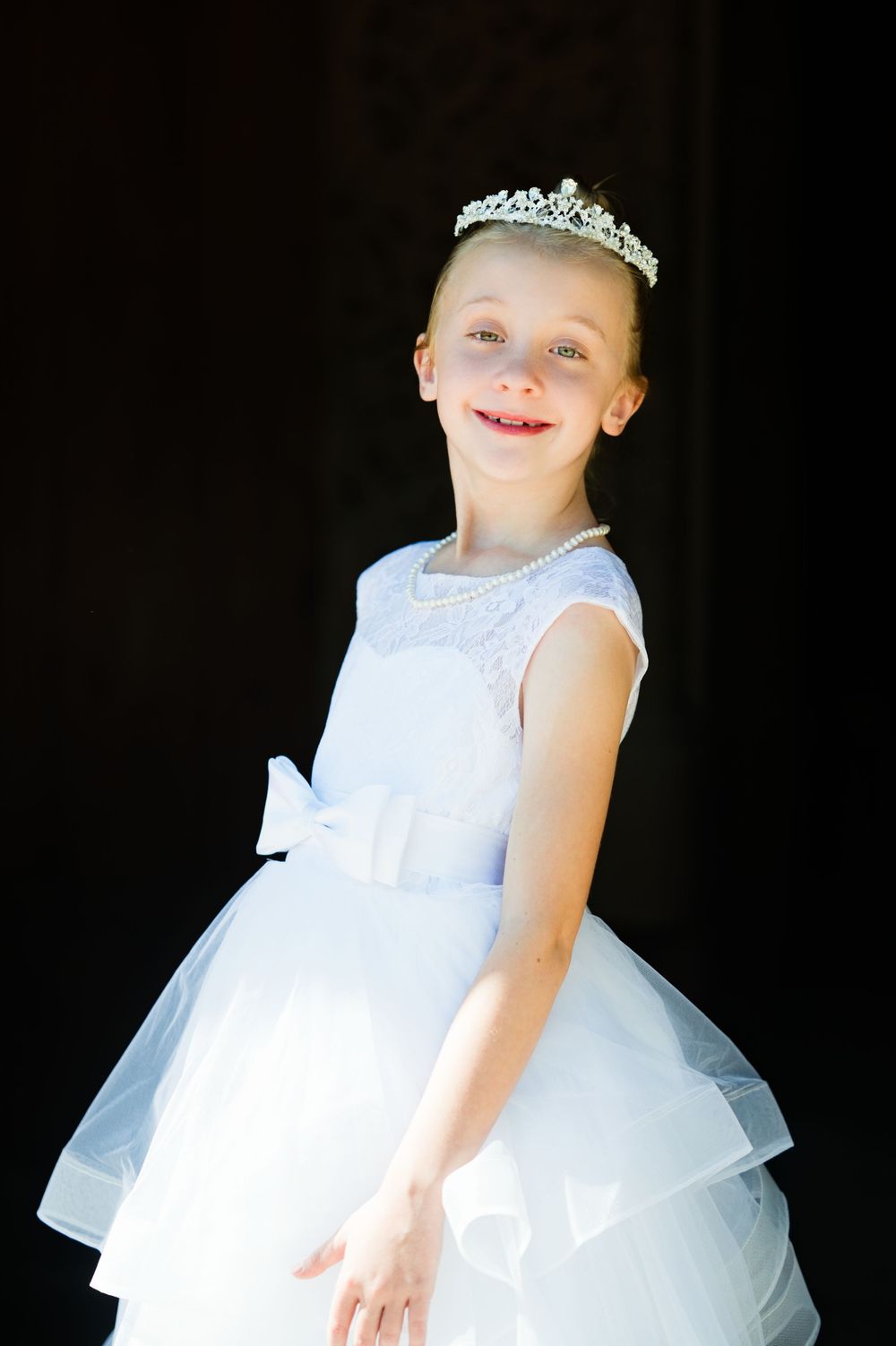 Young girl in white tiara and layered white dress smiling against dark background.