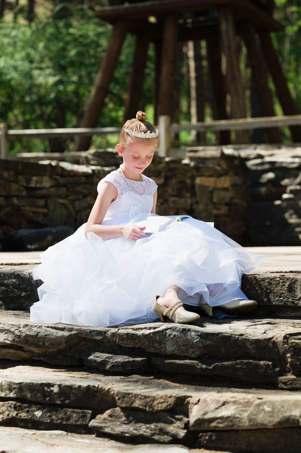 Young girl in white ball gown and tiara sits on stone steps outdoors, looking down gracefully.