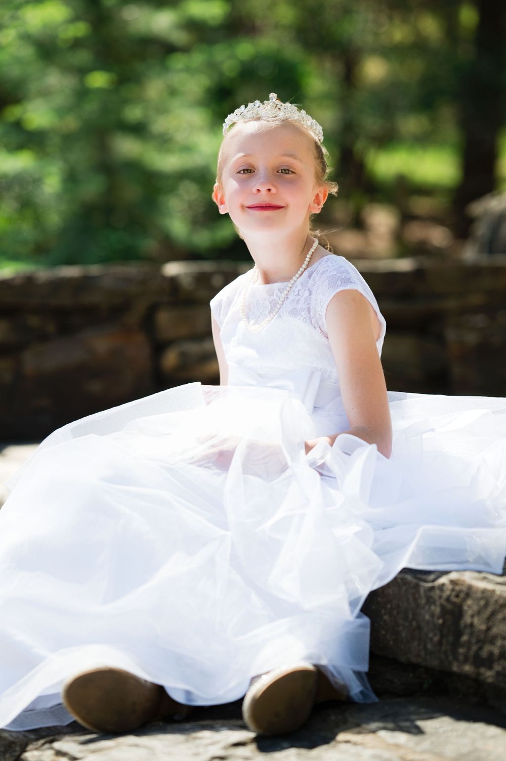Young girl in white First Communion dress and tiara sits on stone steps outdoors, smiling joyfully.
