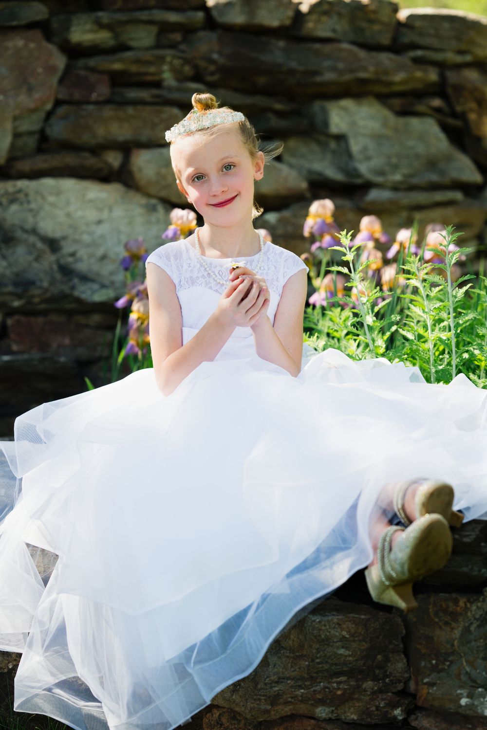 Young girl in white tulle dress sitting on stone wall surrounded by purple flowers and greenery.
