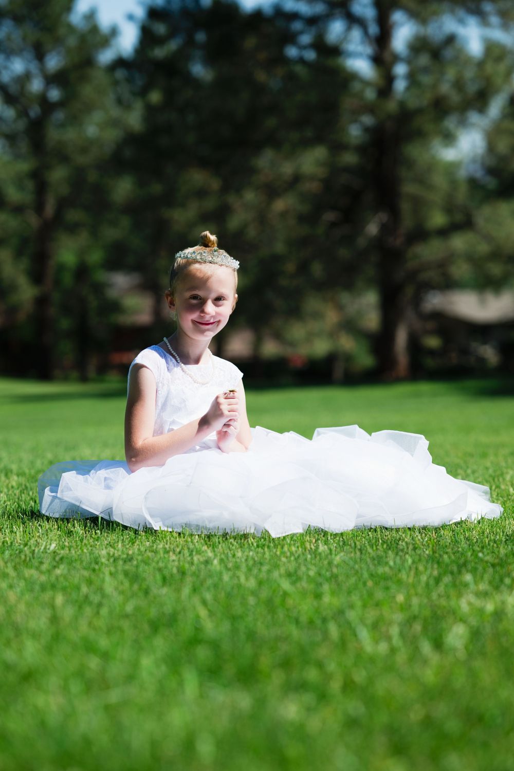 A young girl in a white tulle dress and tiara sits smiling on green grass outdoors.