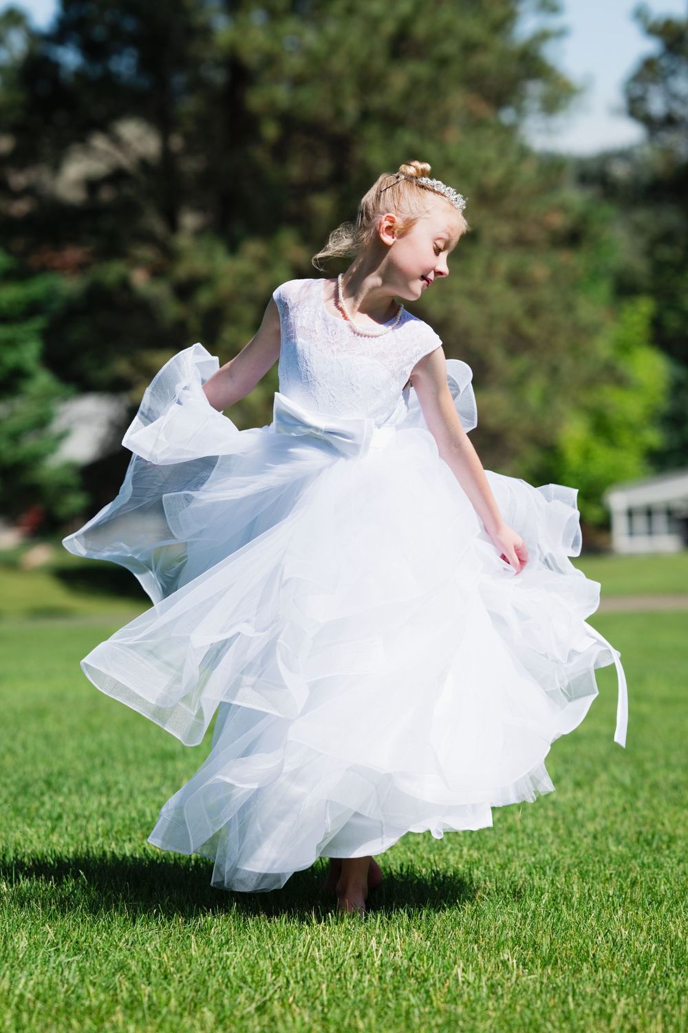 Young girl spinning in white tulle dress on green lawn in sunny park.
