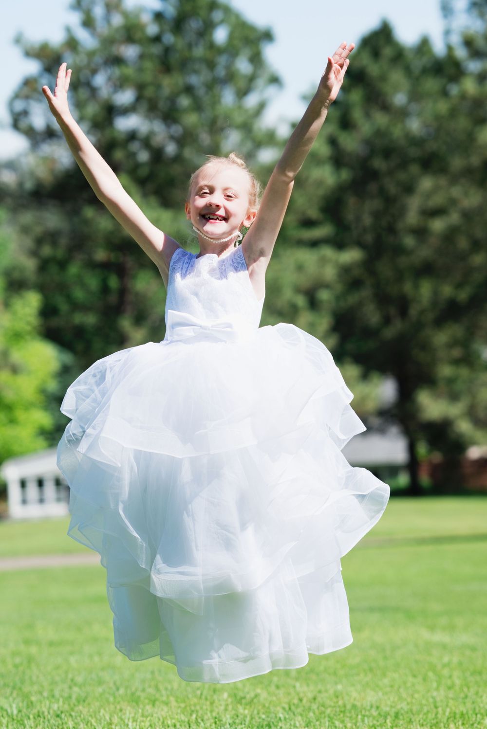 A joyful young girl in a white tulle dress jumps with arms raised on a sunny green lawn.