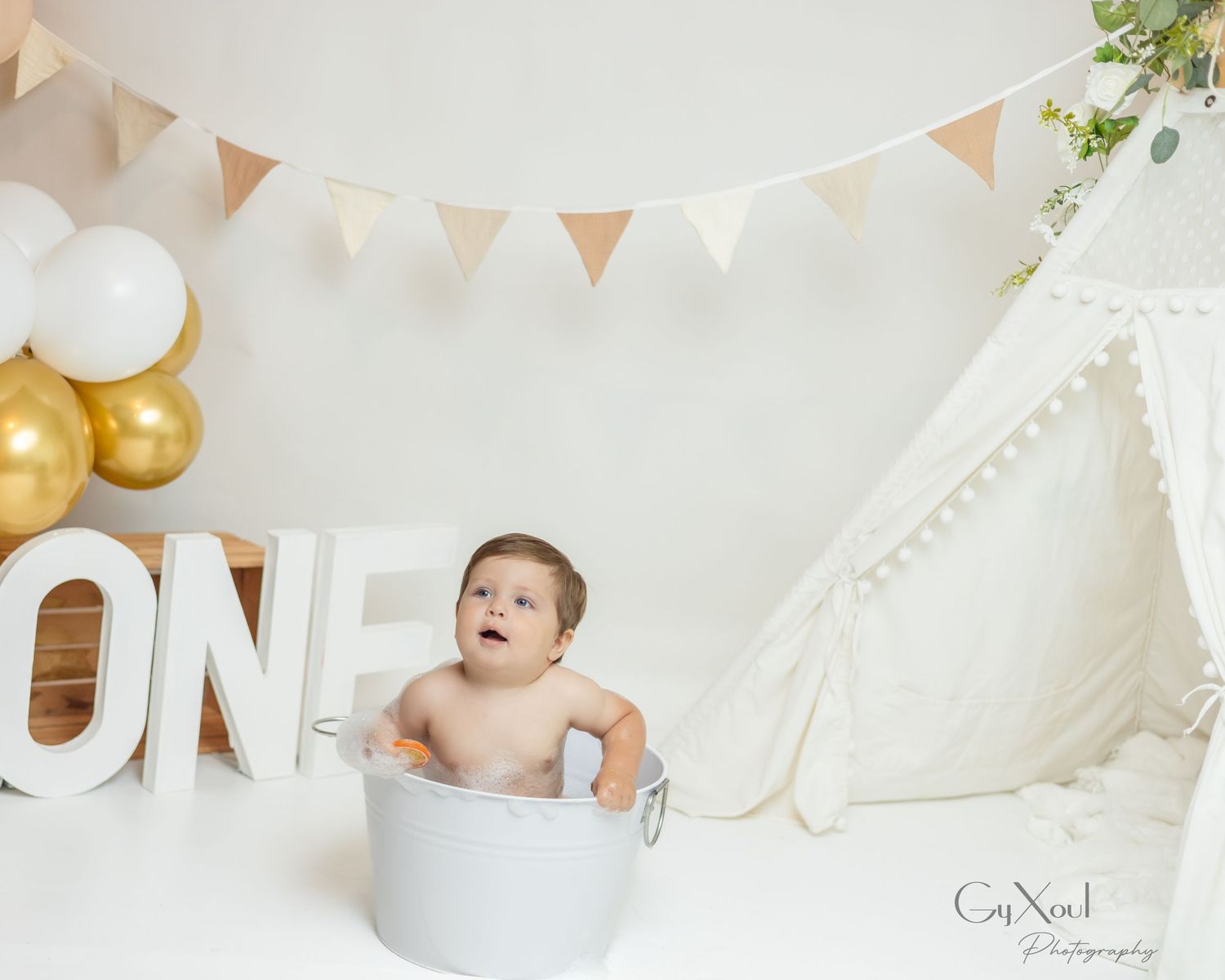 Baby sits in a tin tub beside ONE letters, a teepee, and gold balloons for a first birthday cake smash photo.