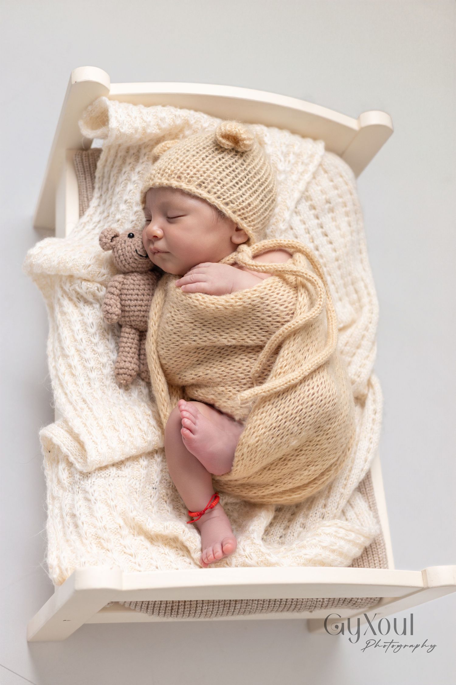 Newborn baby sleeping in tiny white wooden bed, wrapped in beige knit blanket with bear hat and stuffed toy.