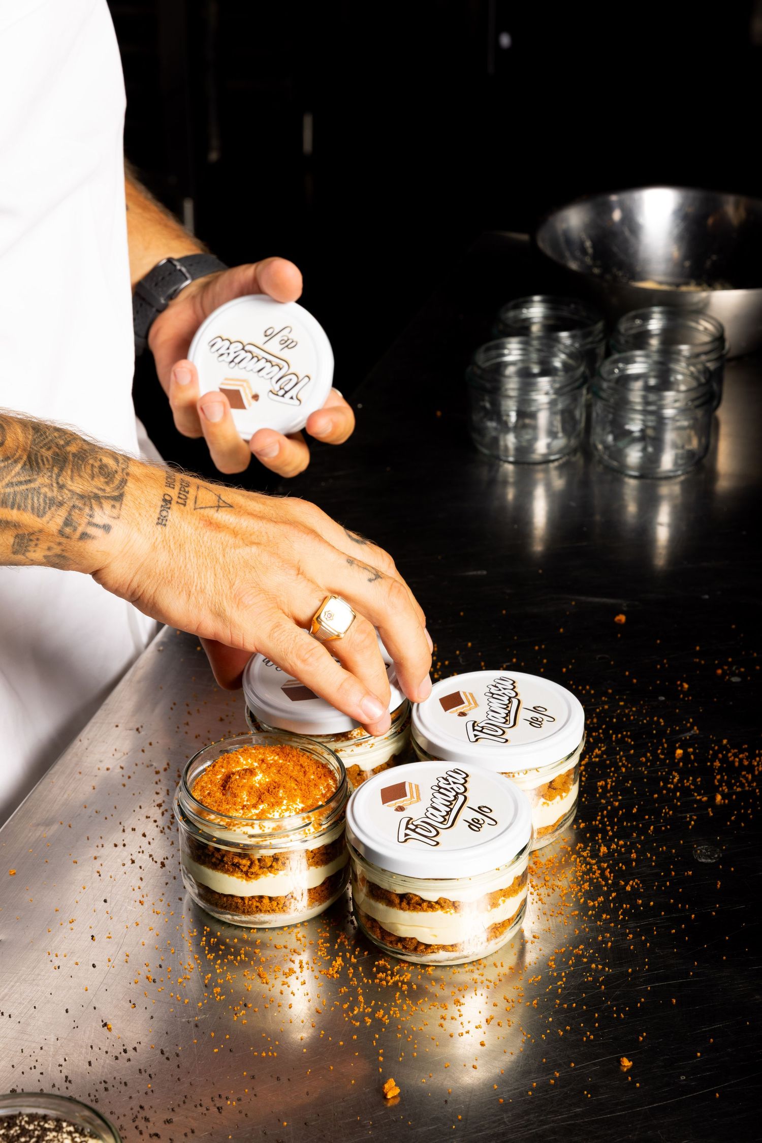 Sequence of hands with tattoos opening small glass jars against a dark tiled background.